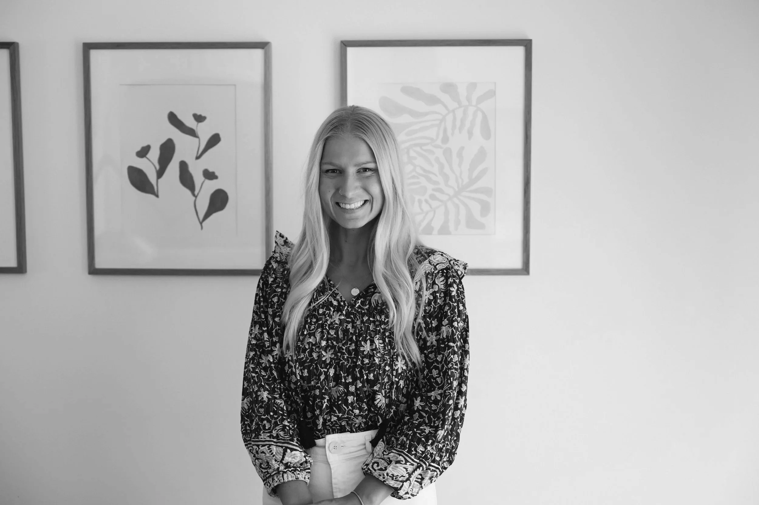 A woman with long, wavy hair smiling and standing in front of a wall with framed botanical artwork.