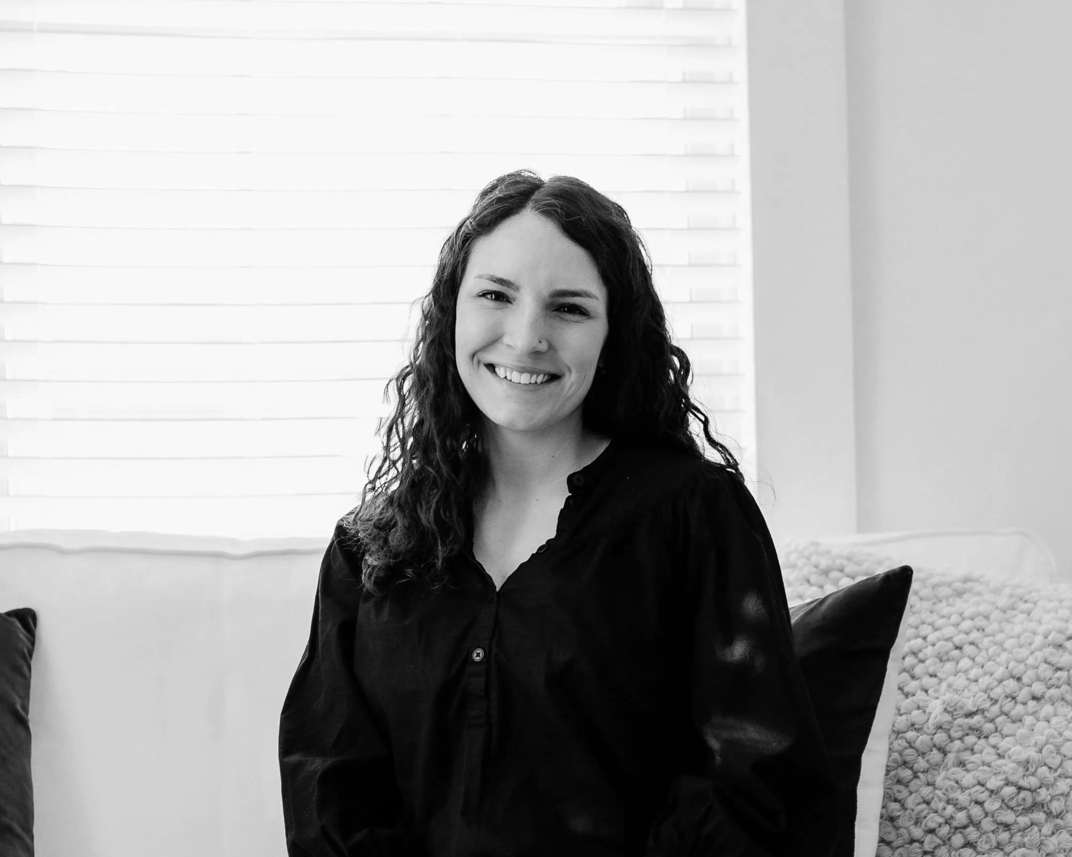 A woman with curly hair smiling, wearing a dark shirt, sitting in a room with white walls, white window blinds, and a bed with a blanket of white stones.