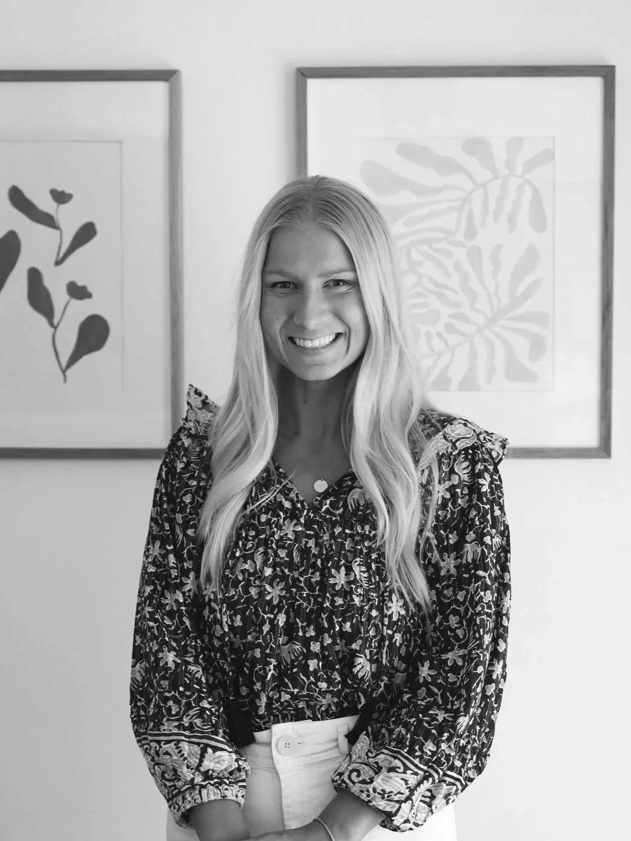 Black and white photo of a smiling woman with long wavy hair standing in front of two framed botanical prints on a wall.