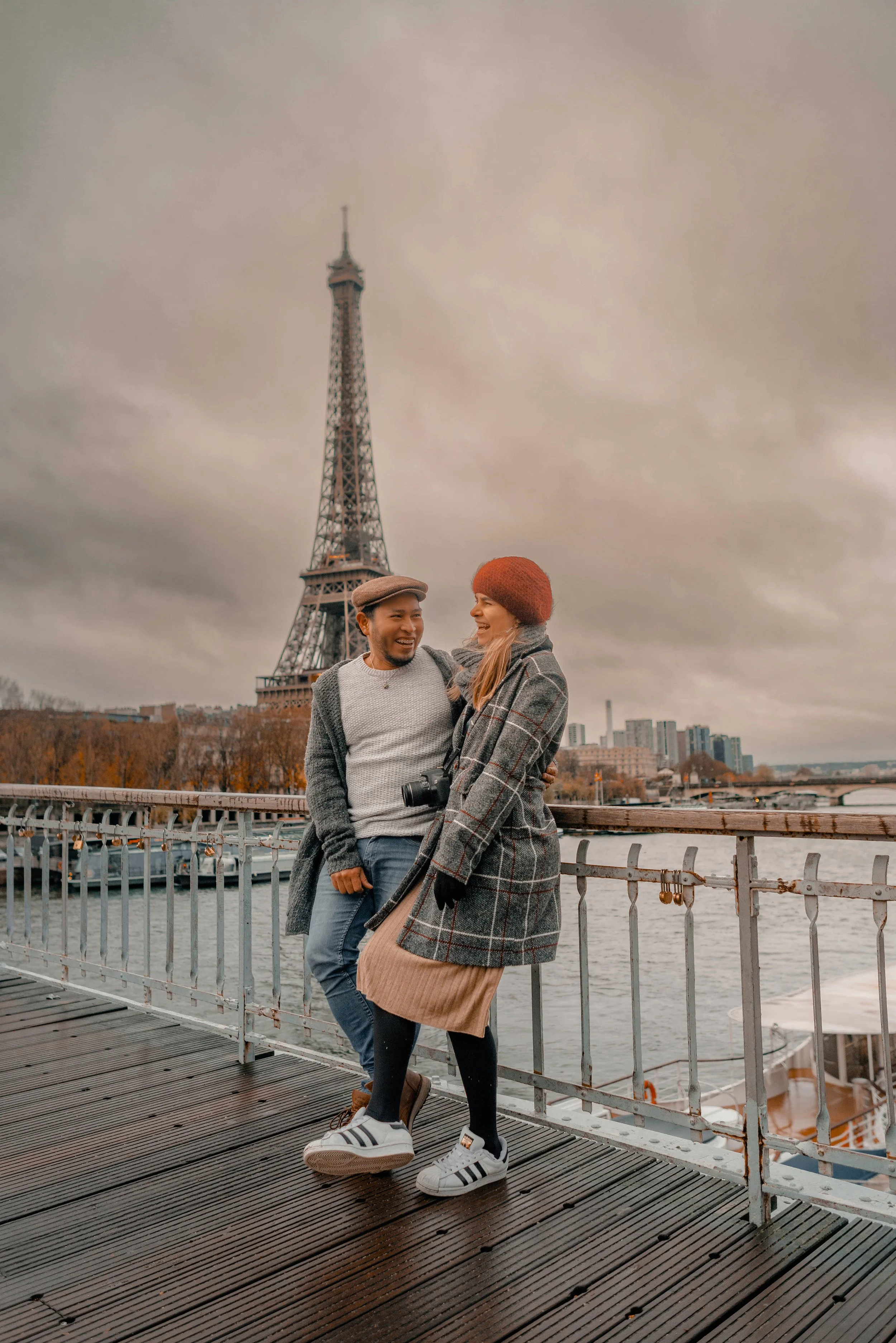 Una pareja sonriendo en un muelle en París, con la Torre Eiffel al fondo y un cielo nublado.