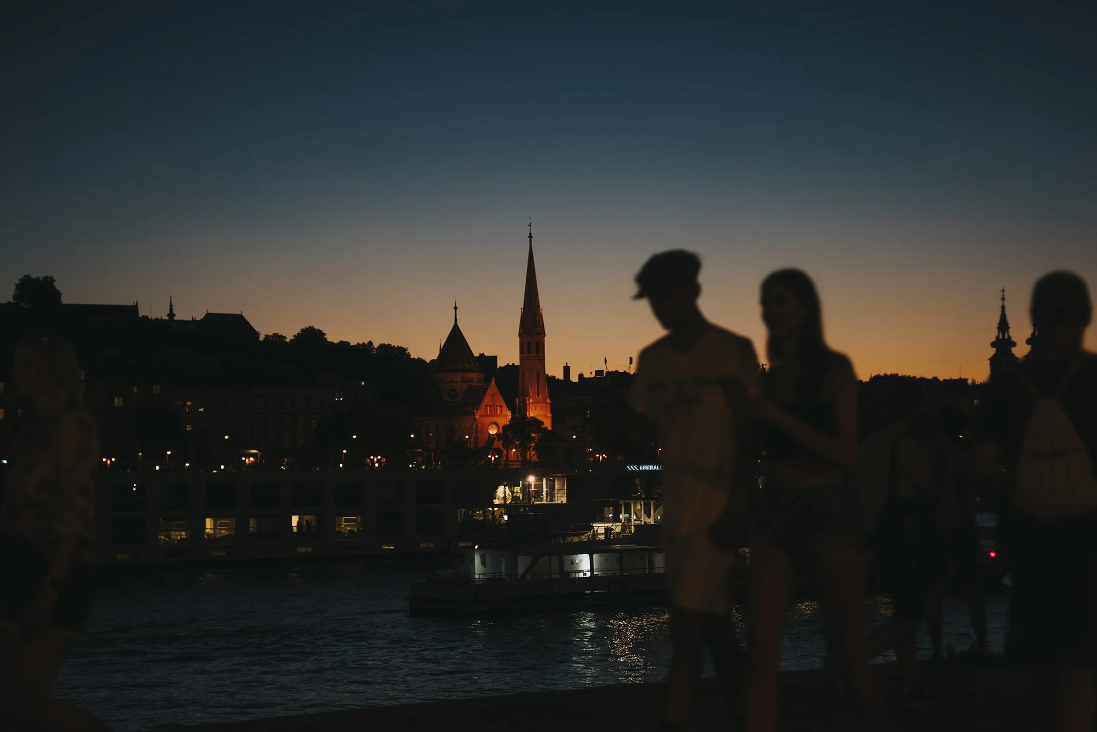 Budapest Silhouettes of people walking near the water at dusk with city buildings and church steeples in the background.