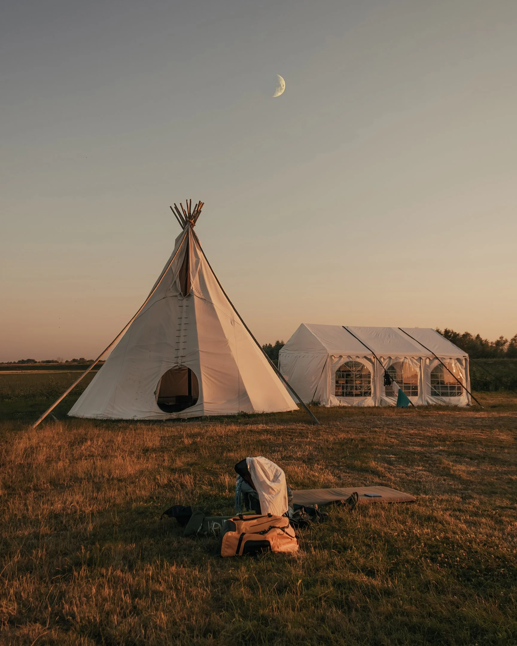 A campsite with a tipis and a tent on a grassy field during sunset, with the moon visible in the sky.