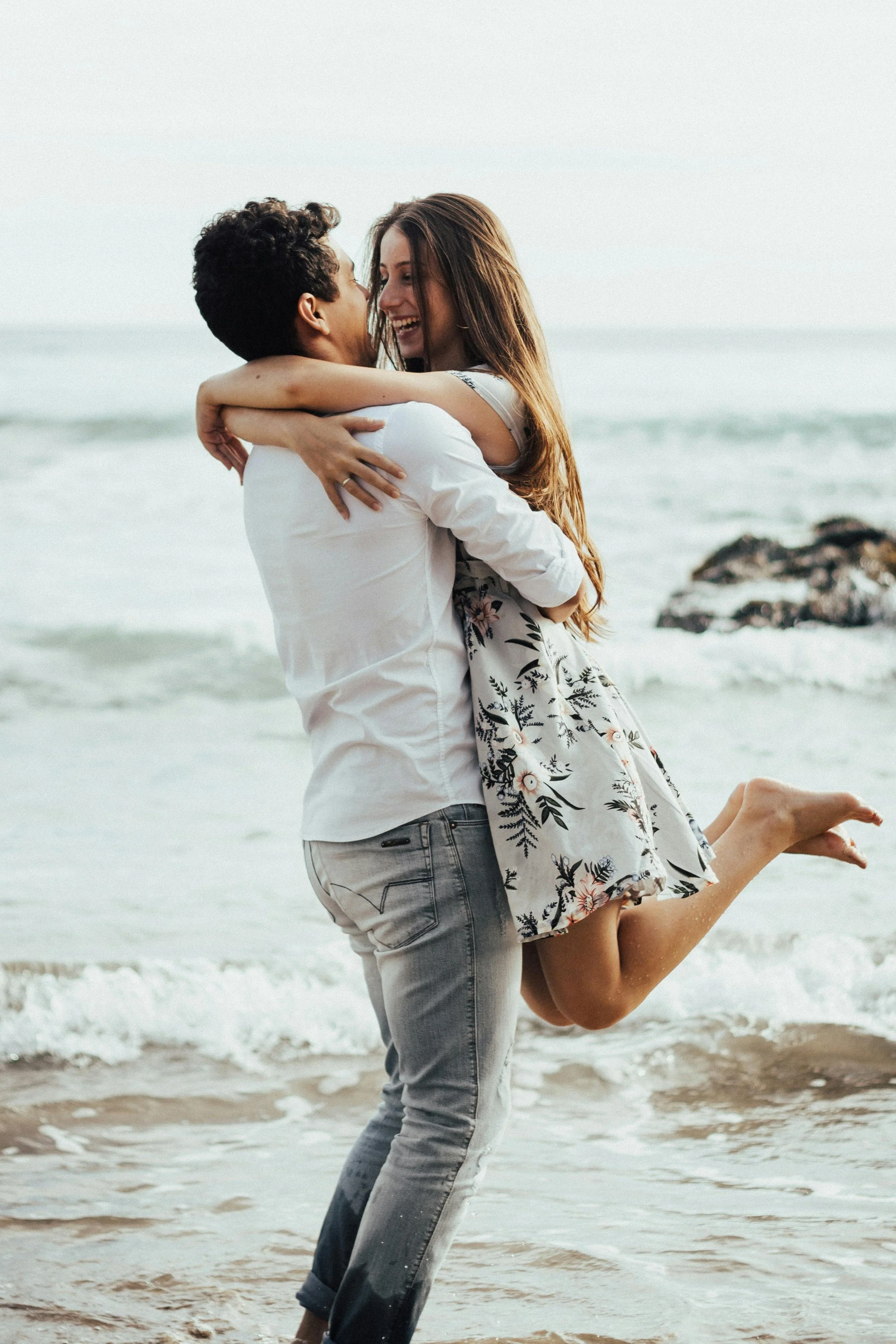 Una pareja feliz abrazándose en la playa, con olas y rocas en el fondo, ella usando vestido con estampado floral y él en camisa blanca y jeans