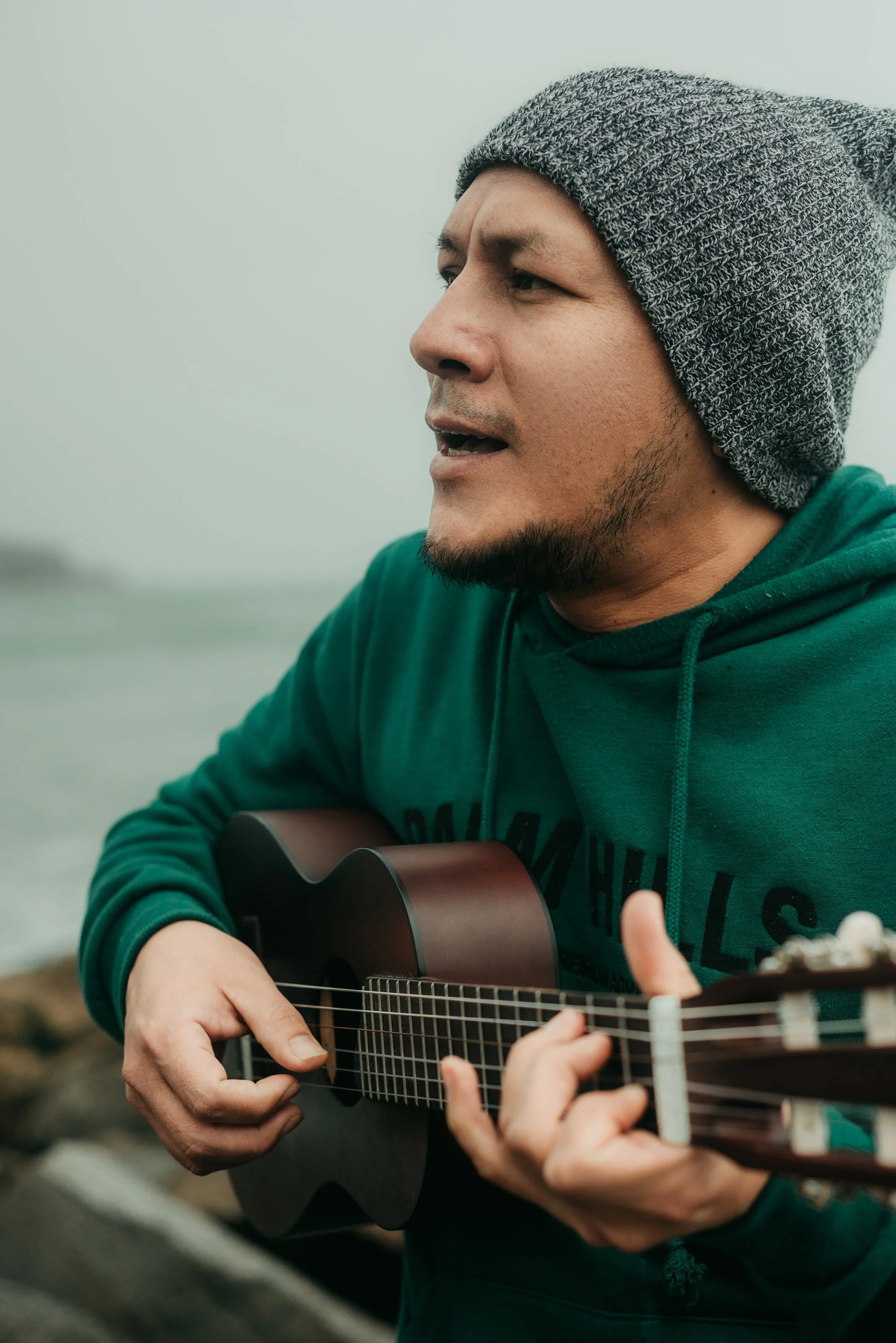 A man wearing a gray knit beanie and a green hoodie playing an acoustic guitar outdoors near the water, with a cloudy sky in the background.
