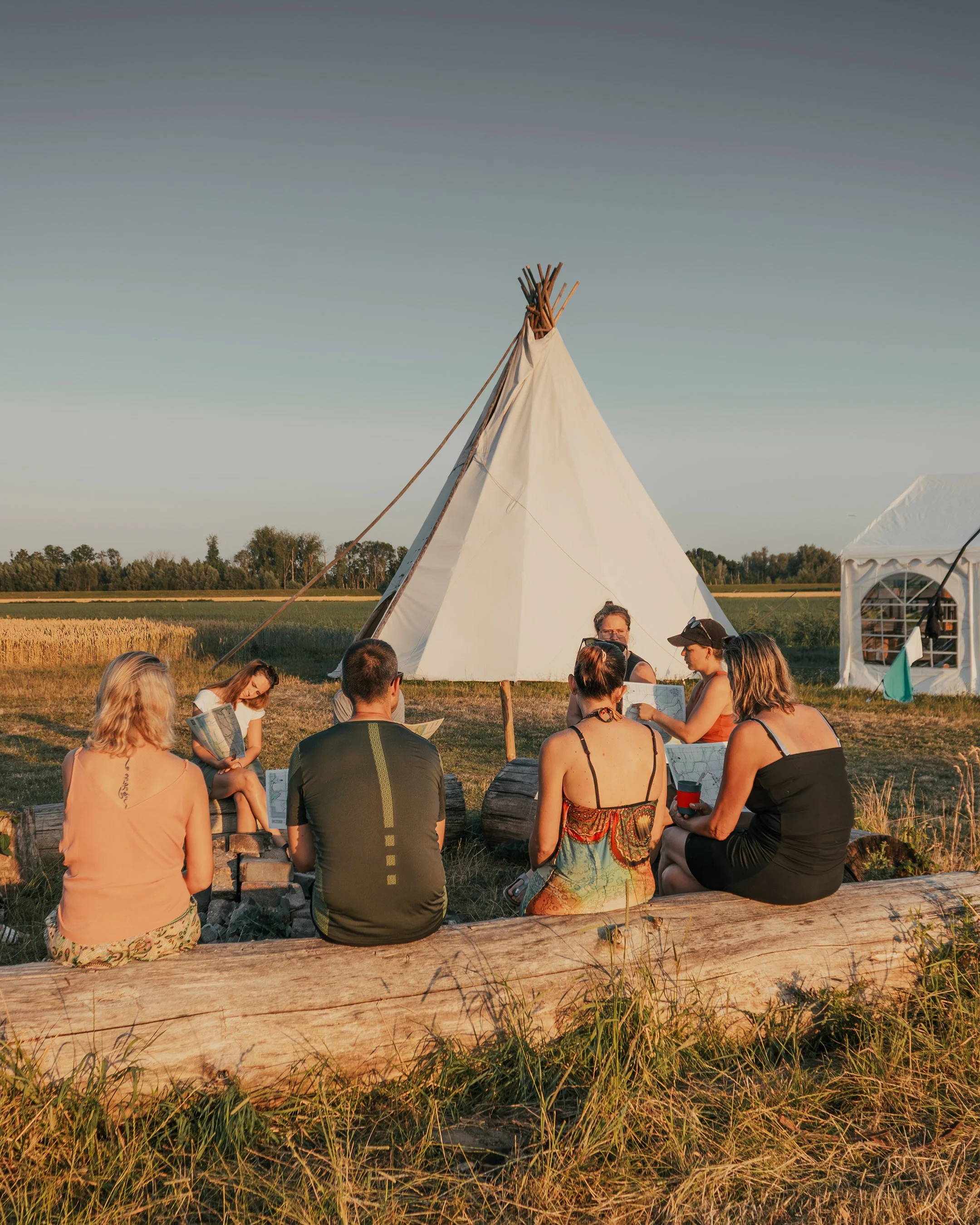 People sitting in a circle around a campfire near a large white tipi tent on open field during sunset.