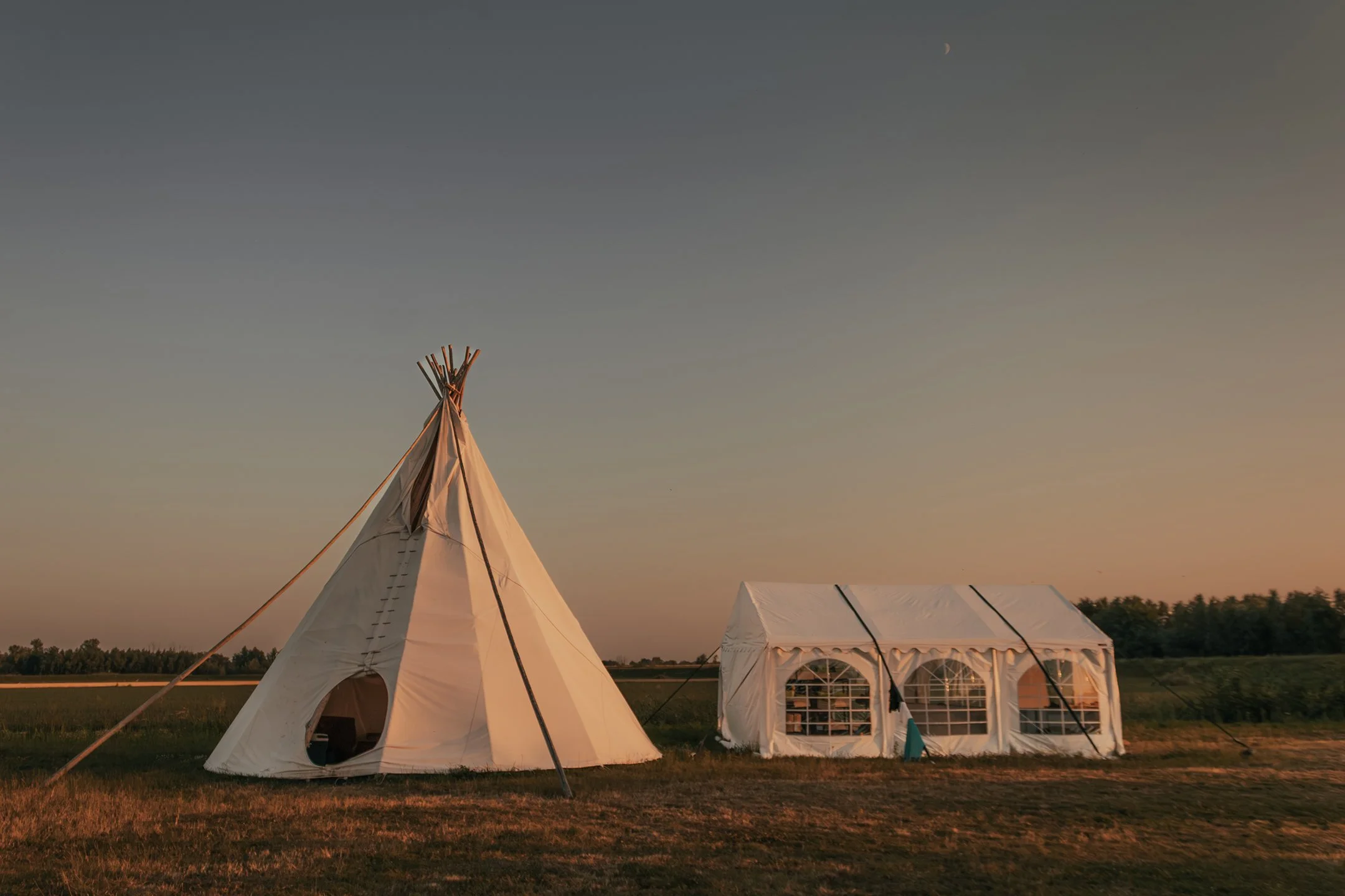 A tipi tent and a white canopy tent are set up in a grassy field during sunset with a clear sky and a small crescent moon.