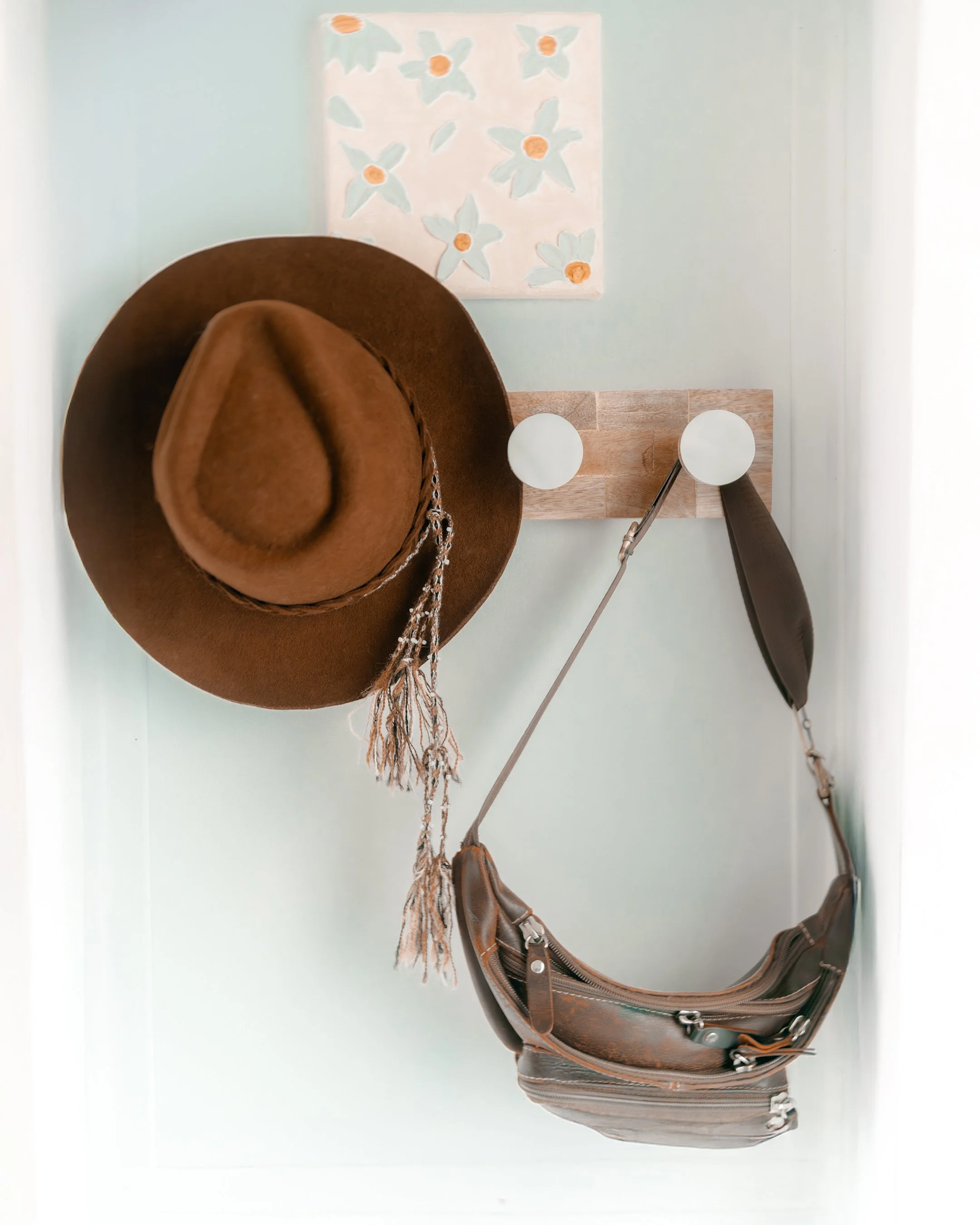 A brown cowboy hat, a leather crossbody bag, and a feather hanging on a wall hook with a light-colored wall and a floral painting in the background.