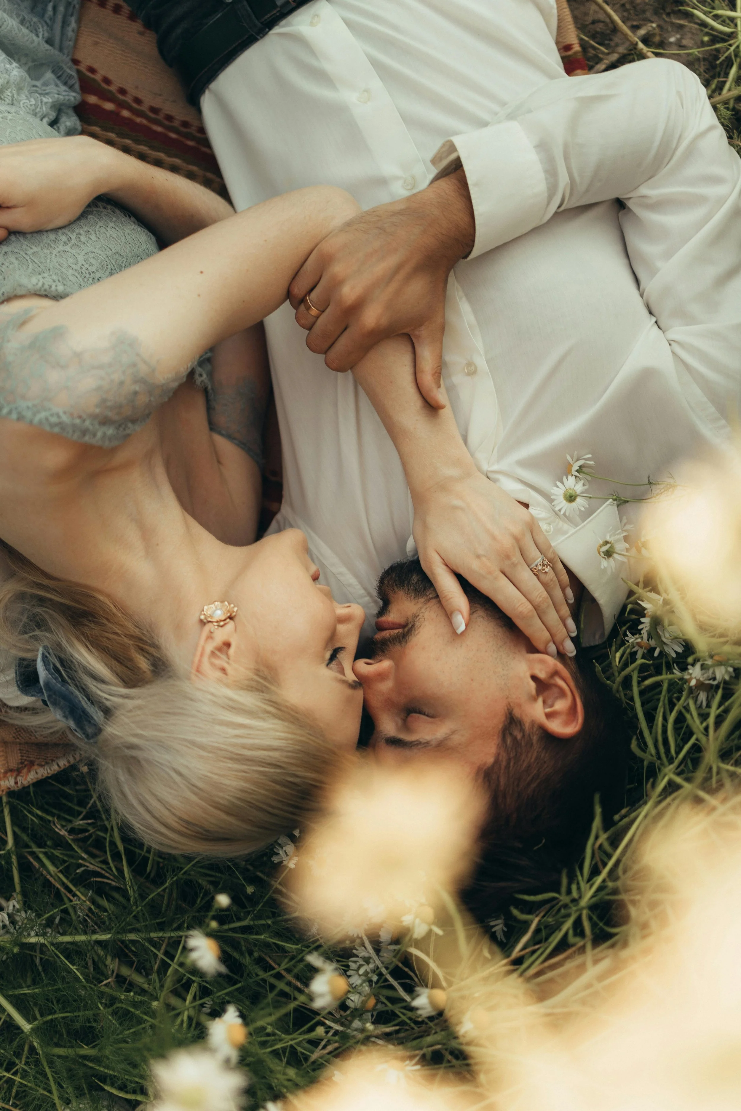 Pareja acostada en flores, ella con oreja con pendiente, él con barba y cabello oscuro, ambos con los ojos cerrados y en un momento íntimo de beso.