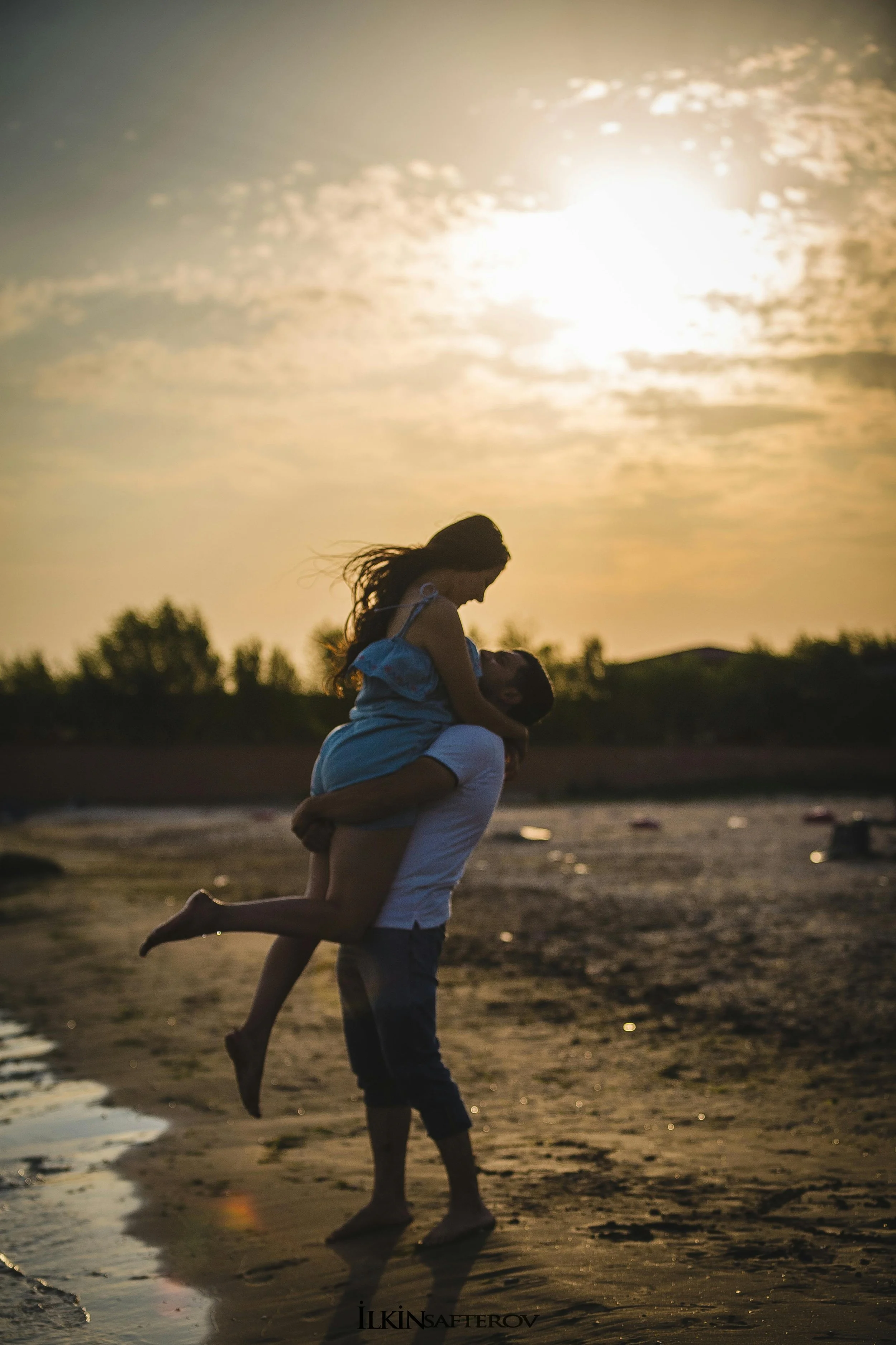 Pareja en la playa, el hombre levanta a la mujer mientras ella lo mira. Atardecer con cielo nublado y sol brillante, sombra en la arena húmeda.