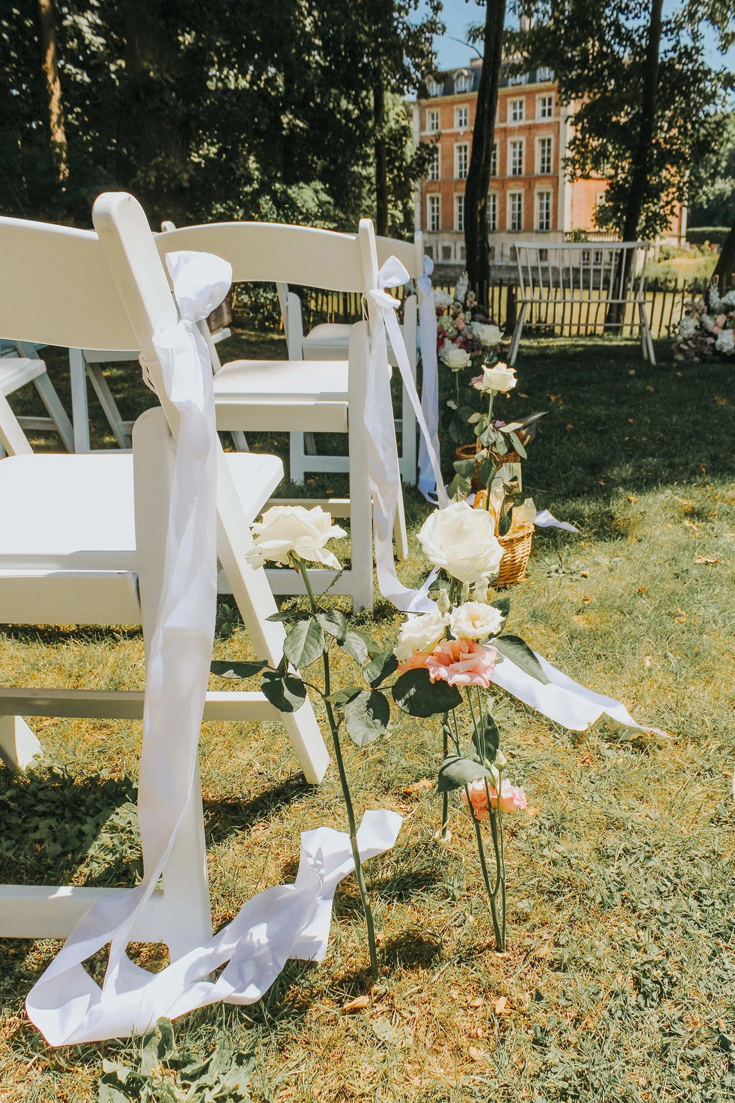 Sillas blancas decoradas con cintas blancas, arreglos de flores en el suelo y un edificio antiguo en el fondo, preparativos para una boda al aire libre.