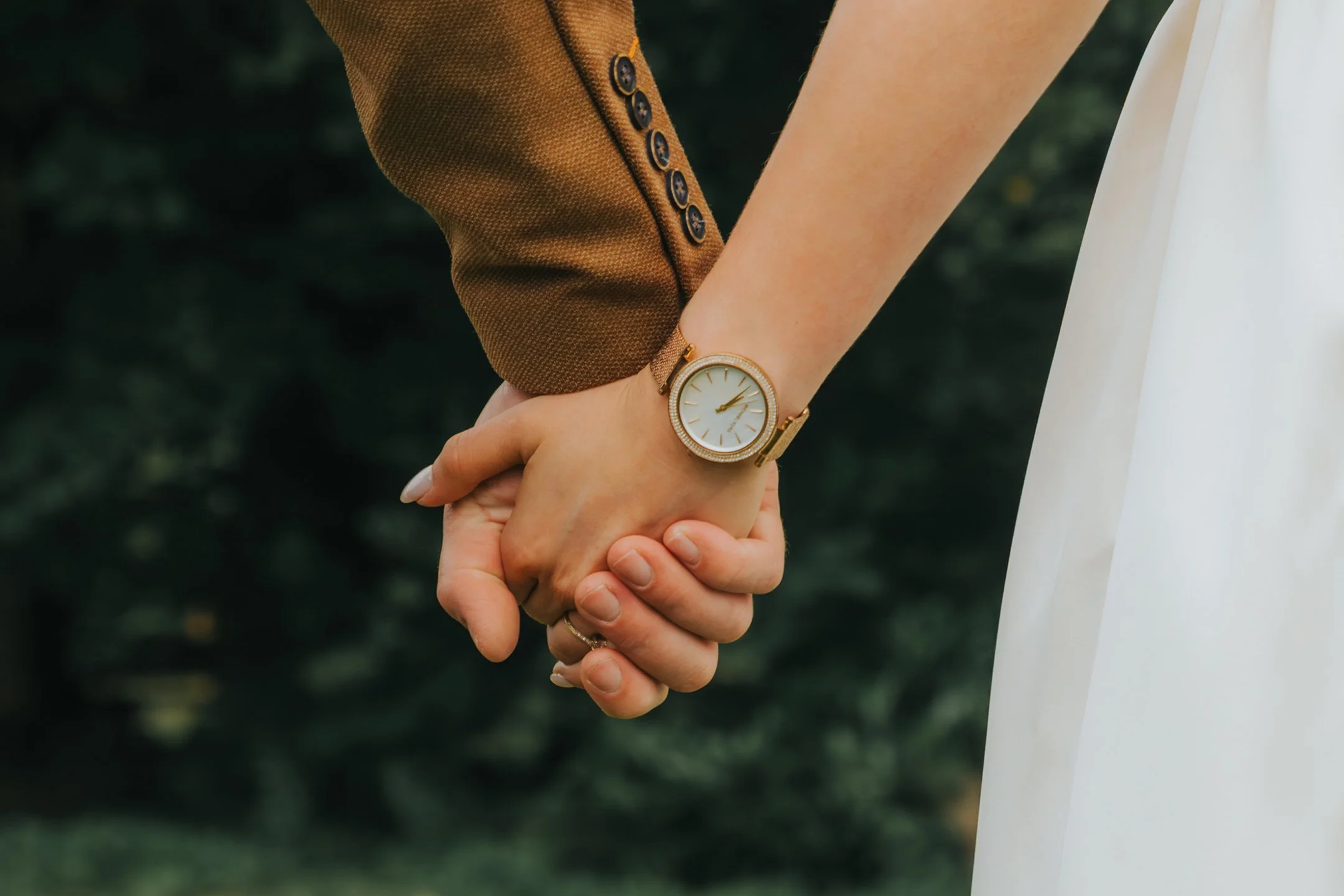 Una pareja que se toma de la mano, llevando puesto un reloj dorado en la muñeca, con fondo de naturaleza verde.