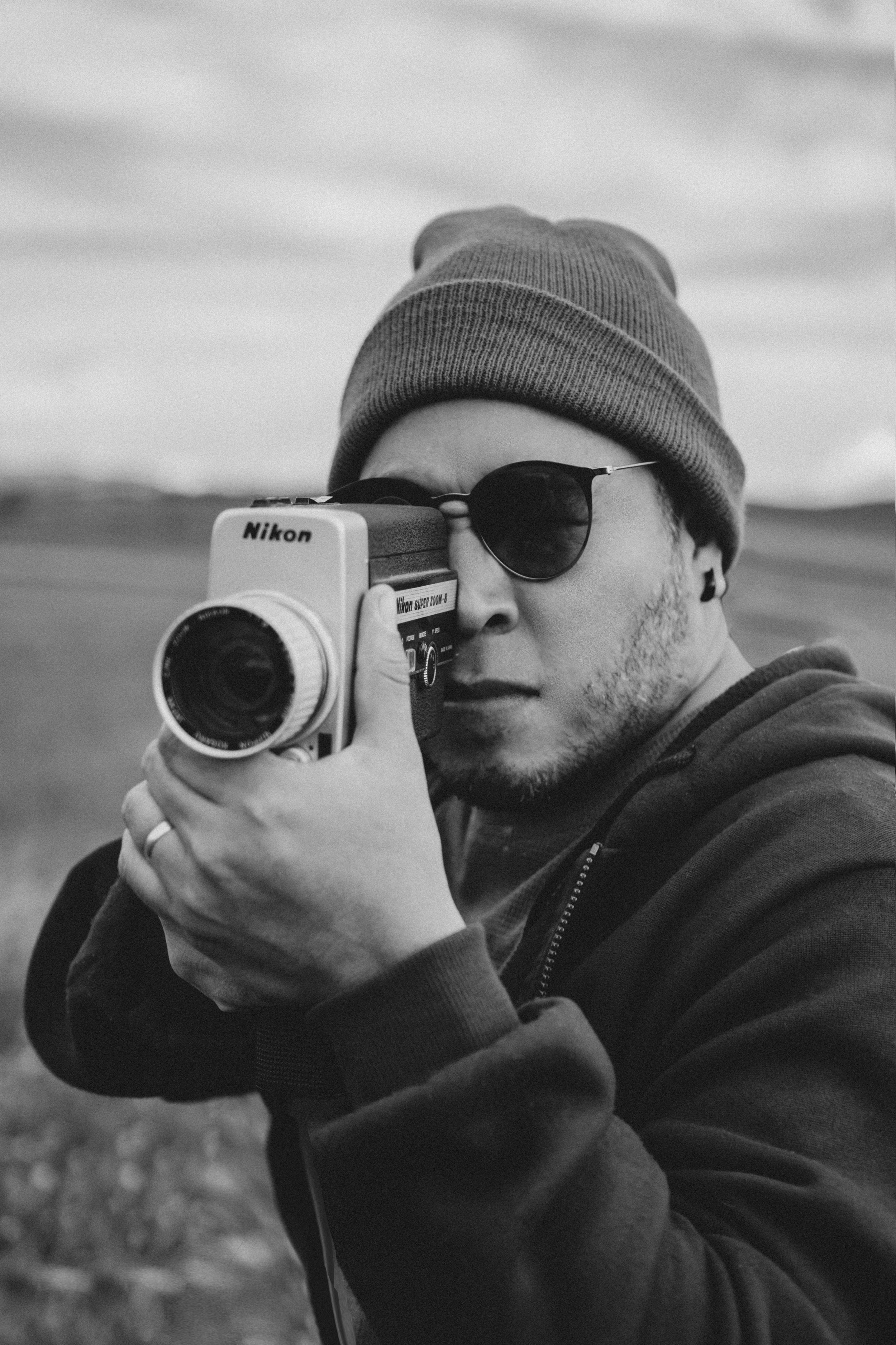 Joven hombre con gorra y gafas de sol sosteniendo una cámara Nikon en un paisaje exterior en blanco y negro.