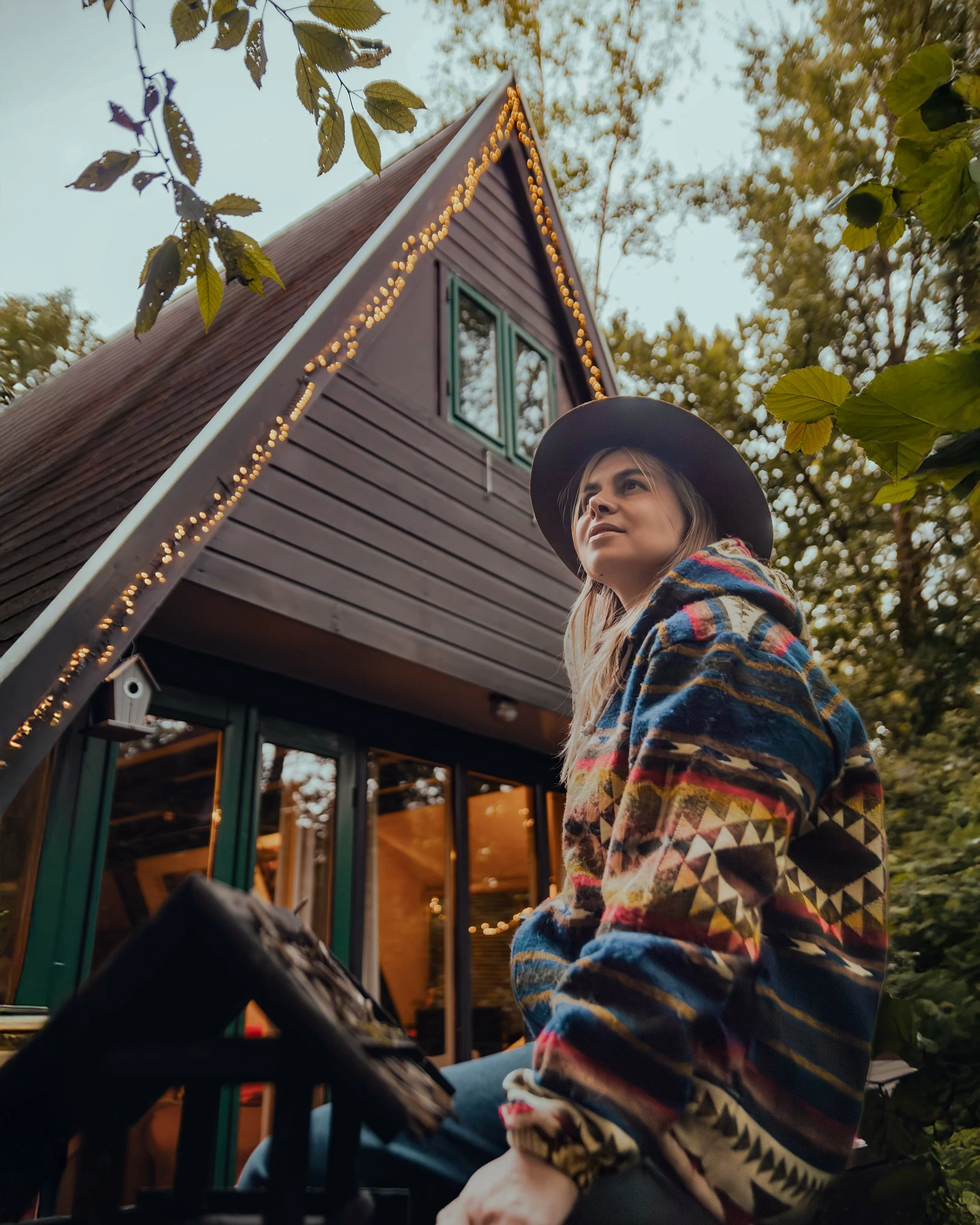 A woman wearing a patterned jacket and hat sitting outdoors near a wooden house with string lights, surrounded by trees.