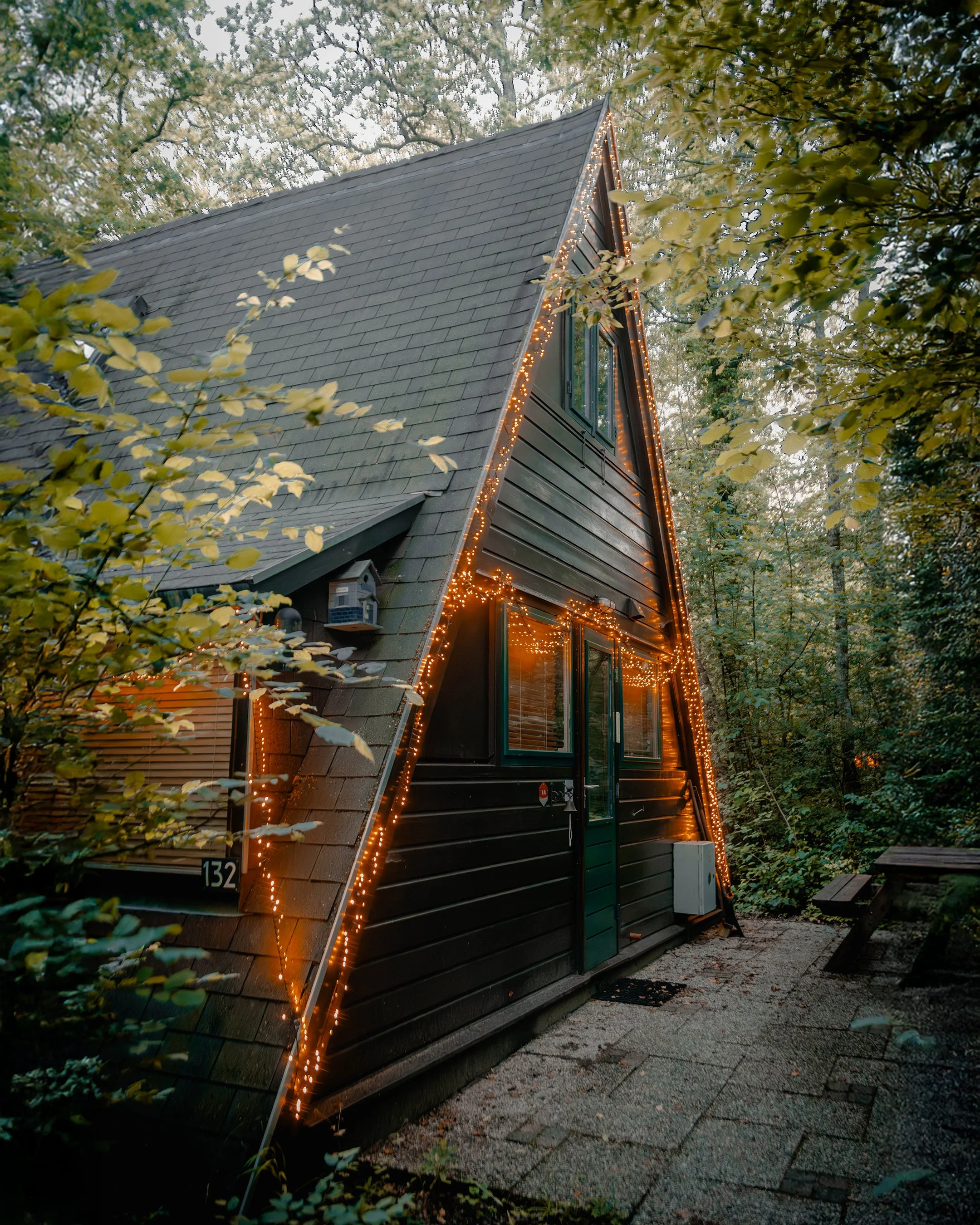 An A-frame cabin decorated with string lights, situated among trees, with a small paved area and benches outside.