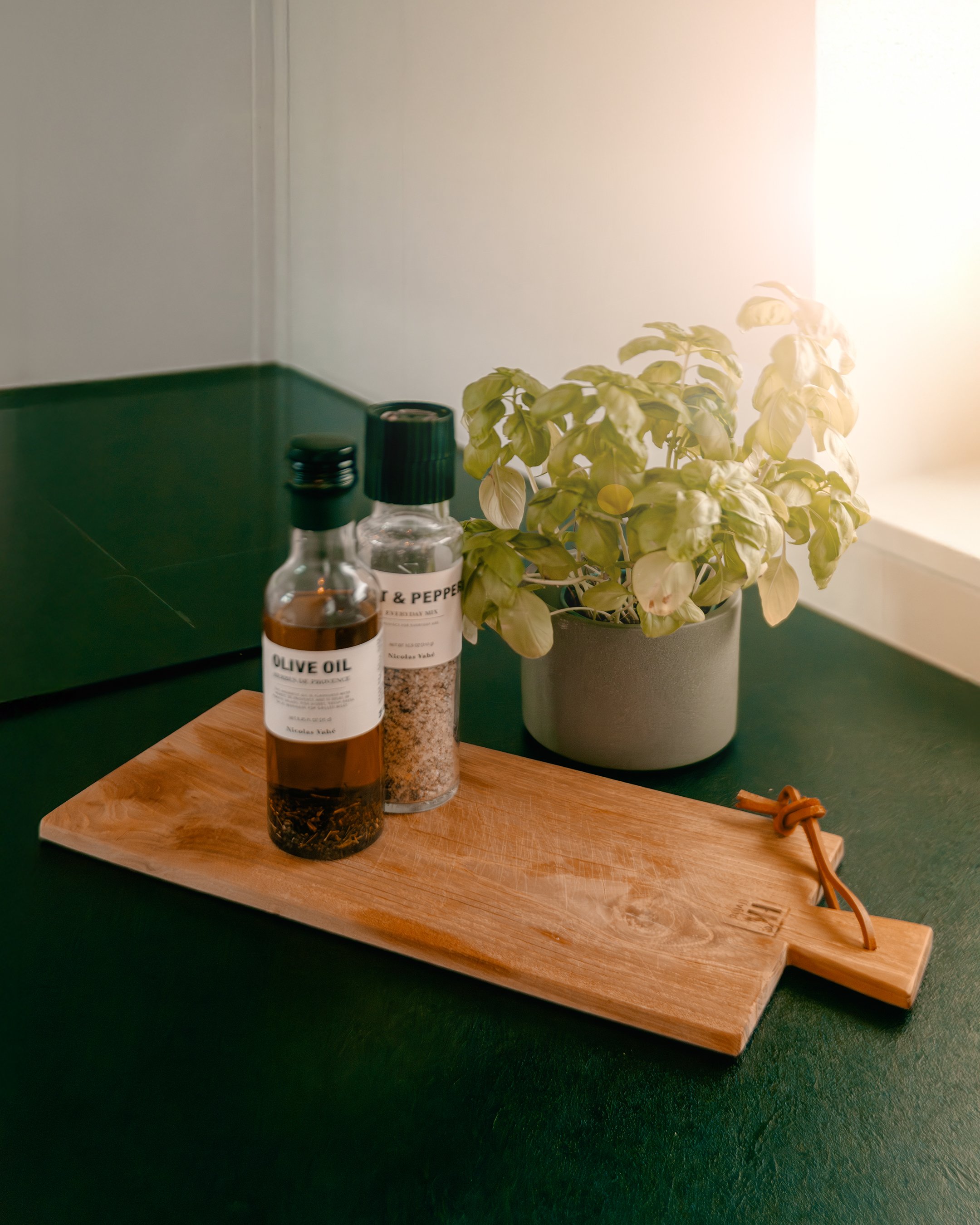 A wooden cutting board with a clip on the handle, placed on a dark green surface. On the board, there are two spice bottles labeled olive oil and salt & pepper. Next to the board, there is a potted plant with green and white leaves, near a window wit