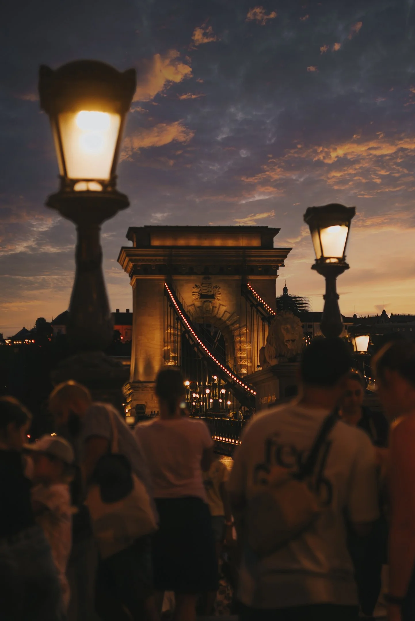 People walking on a bridge at sunset with lit street lamps and a large stone arch in the background.