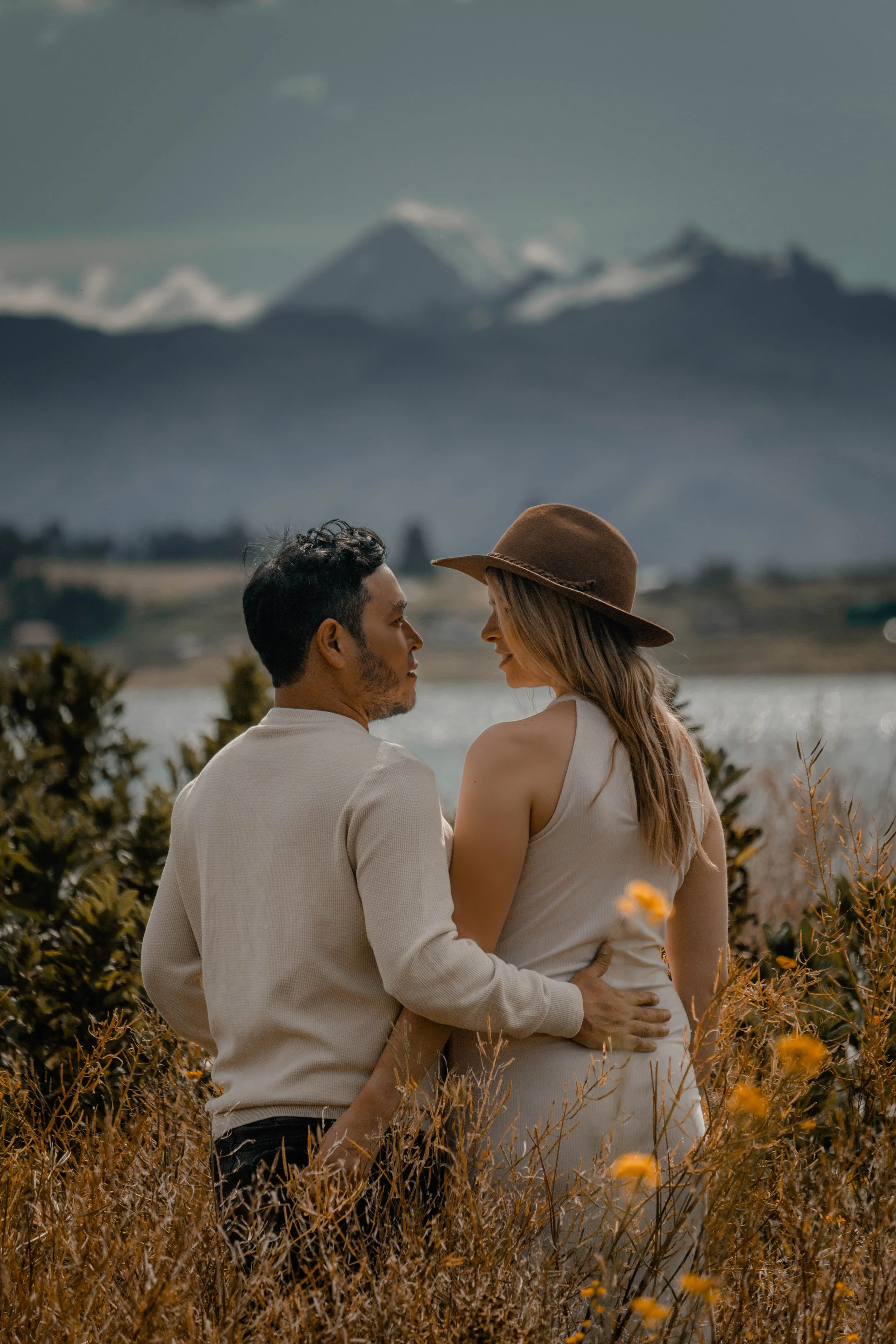 Una pareja observándose frente a un paisaje montañoso con un lago y montañas nevadas en el fondo, rodeados de vegetación.