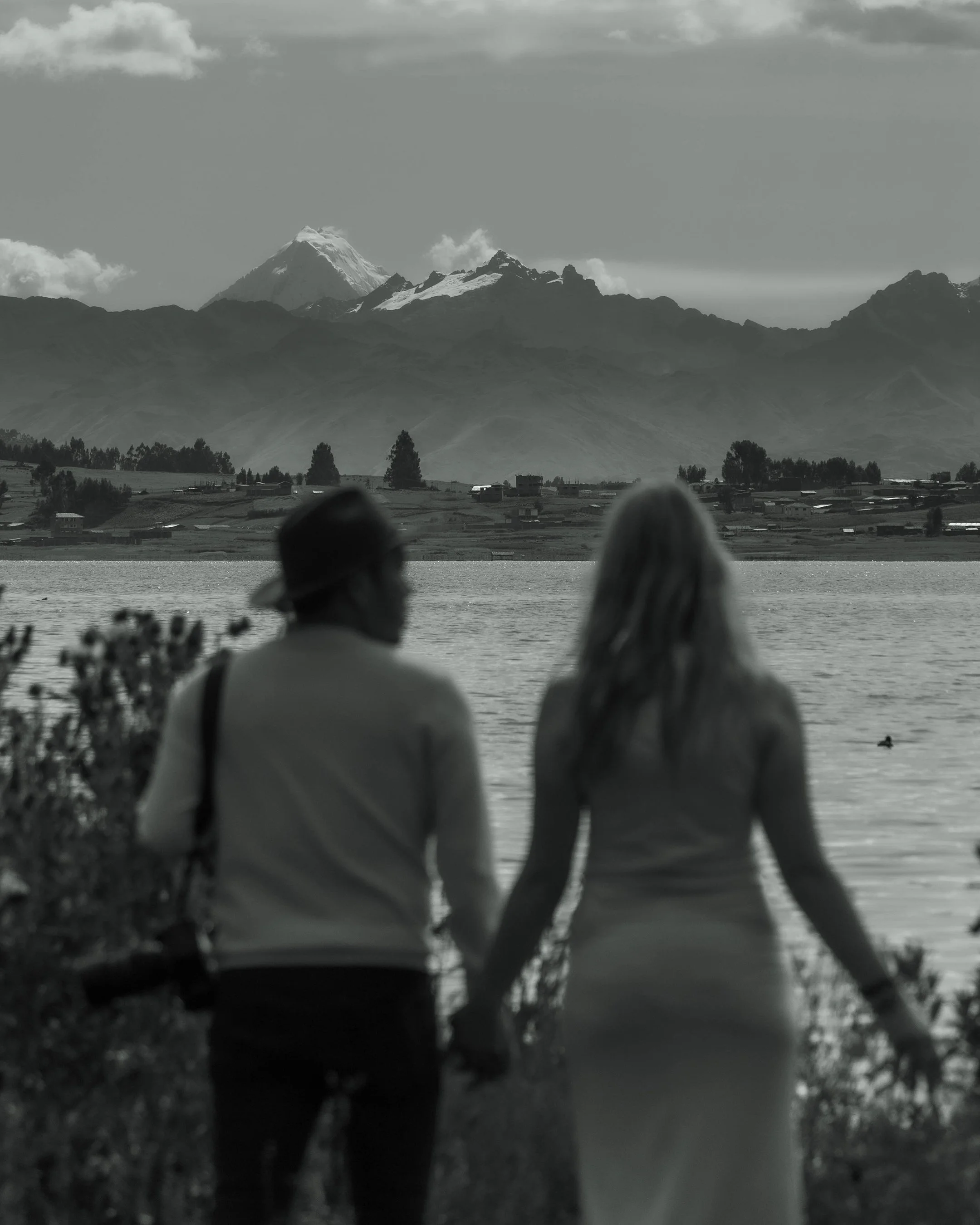 Una pareja sostenido de la mano junto a un lago, con montañas y un volcán en la distancia, en un paisaje en tonos grises.