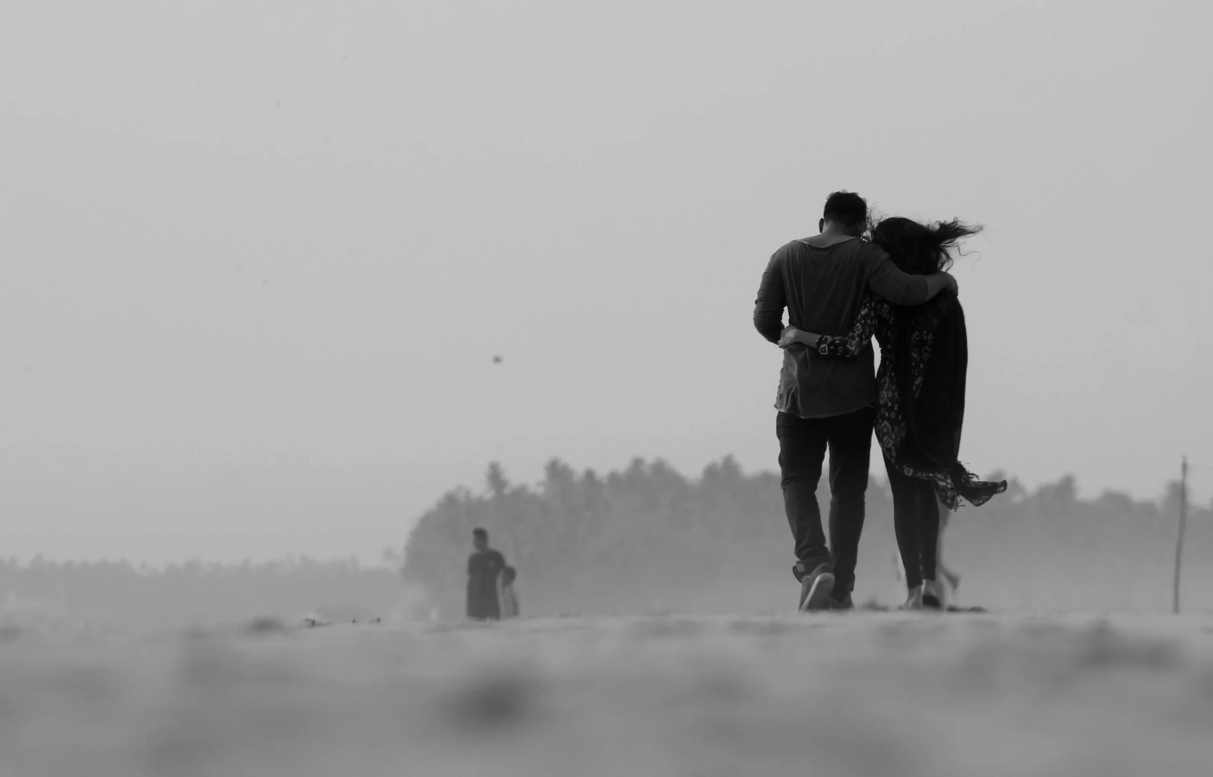 Pareja caminando por la playa, vista de espaldas, en un día nublado, con otra pareja en la distancia