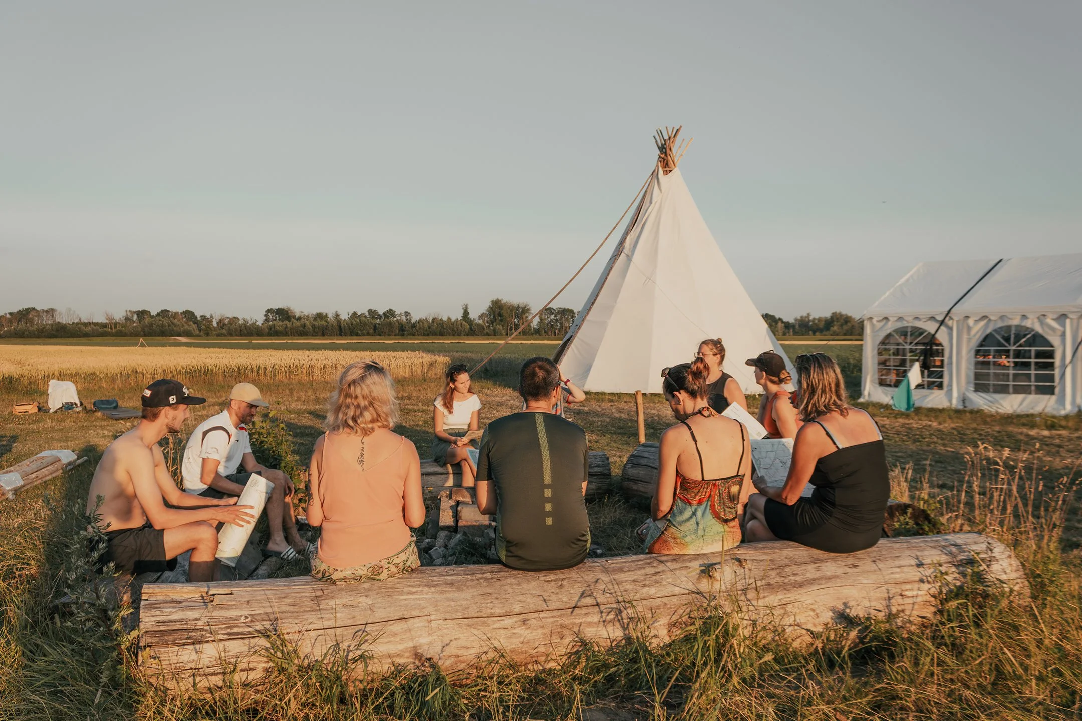 A group of people sitting on logs in a circle outdoors near a teepee and a white marquee tent during sunset, with fields and a clear sky in the background.