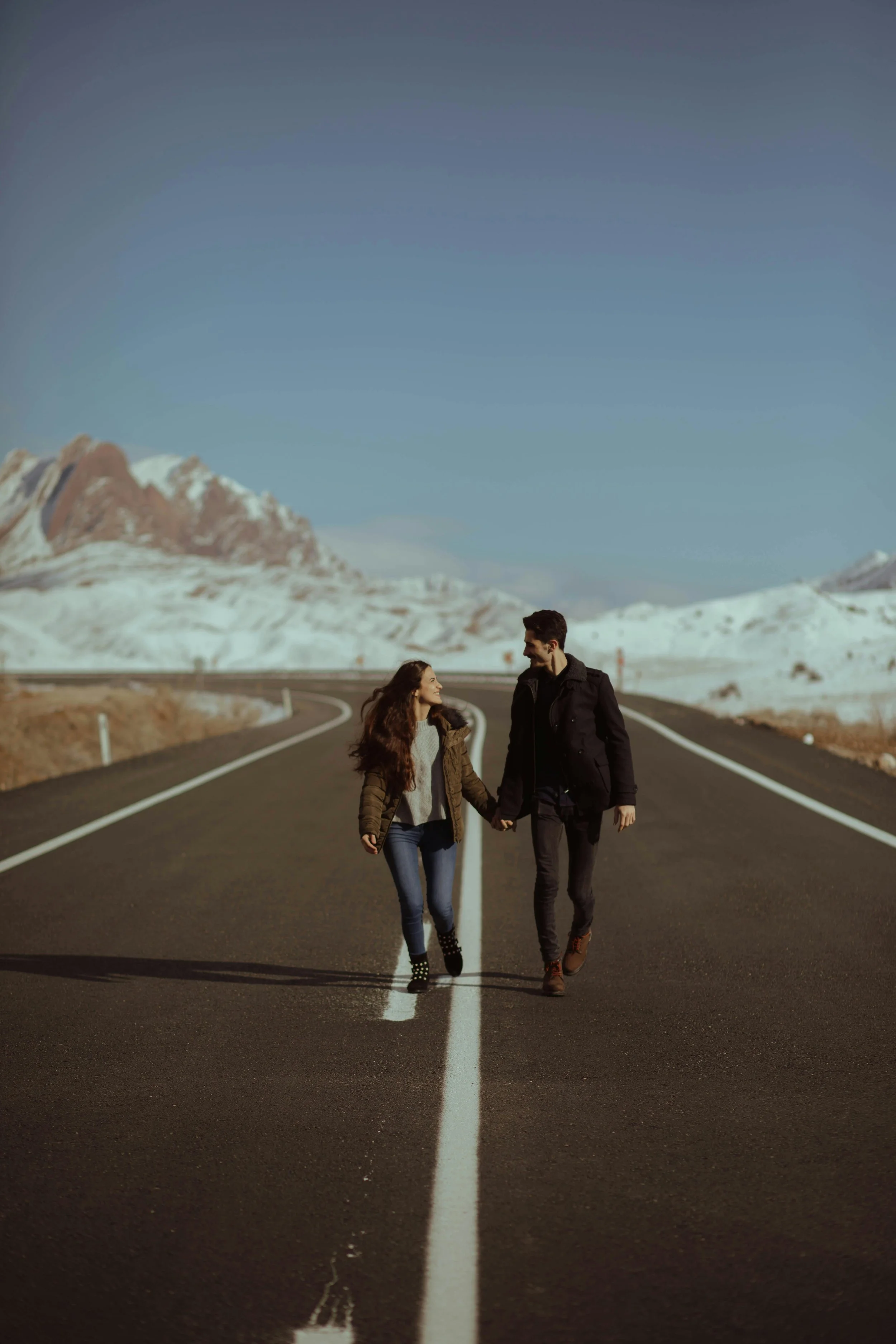 Una pareja caminando junta en un camino de asfalto en un paisaje montañoso con nieve en el fondo