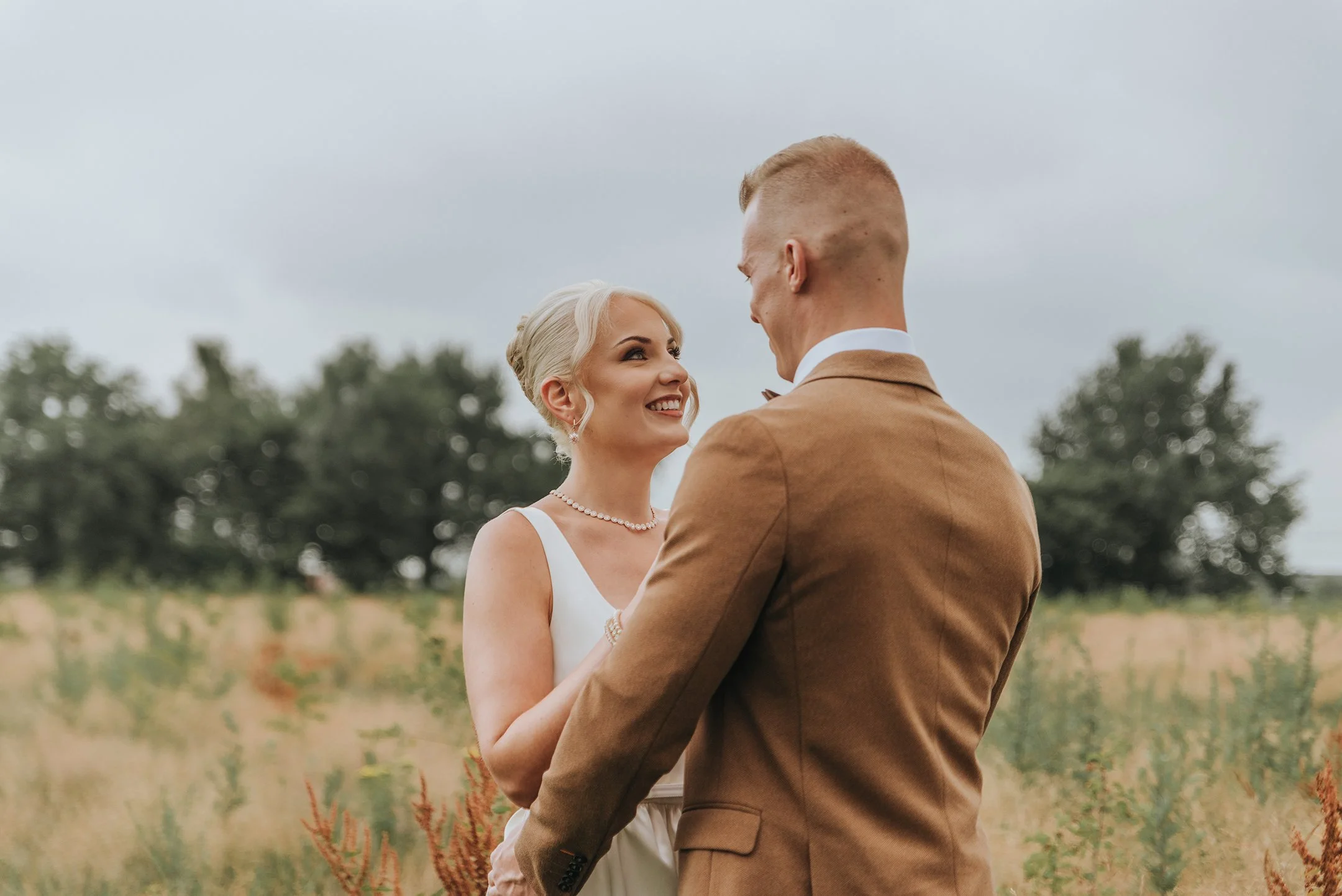 Una pareja de novios en un campo, la mujer viste vestido blanco y perlas, el hombre lleva saco marrón, se miran y sonríen.