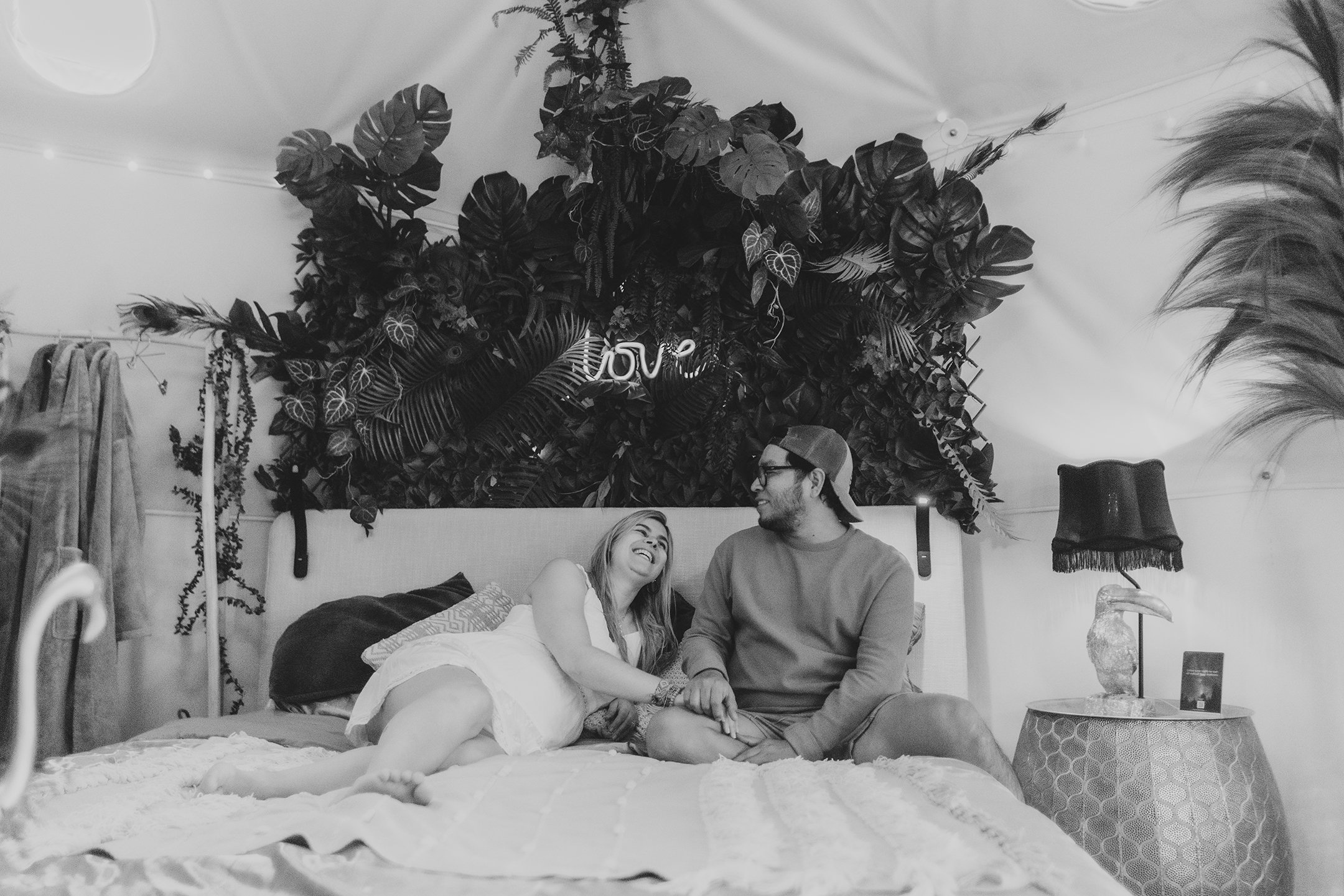 A couple sitting on a bed in a bedroom, laughing and holding hands, with a large decorative plant wall behind them, and a bedside table with a lamp and framed picture on it.