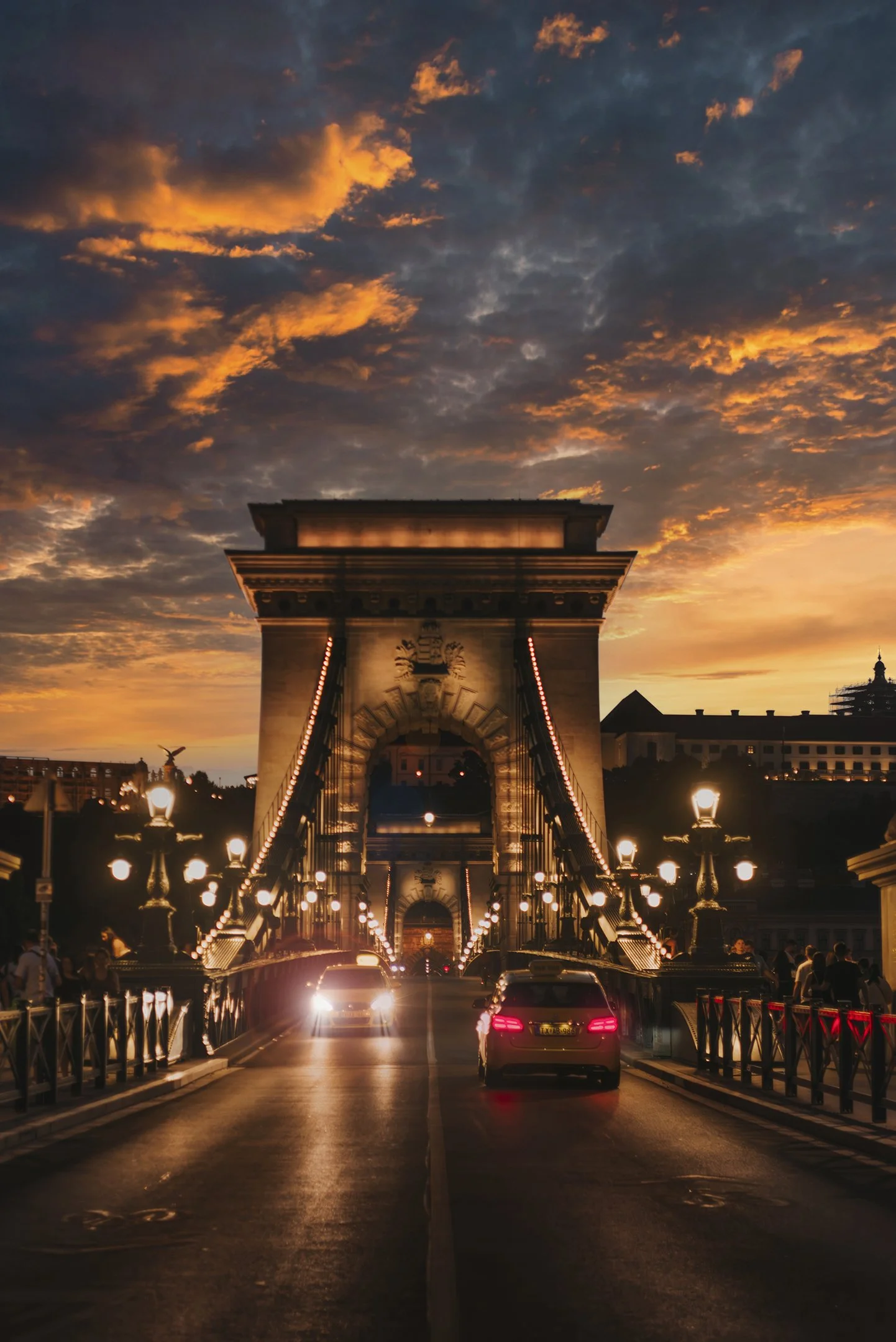 A bridge with illuminated lampposts and cars driving across, leading to a large arch structure, with a dramatic sunset sky filled with orange and dark clouds in the background.