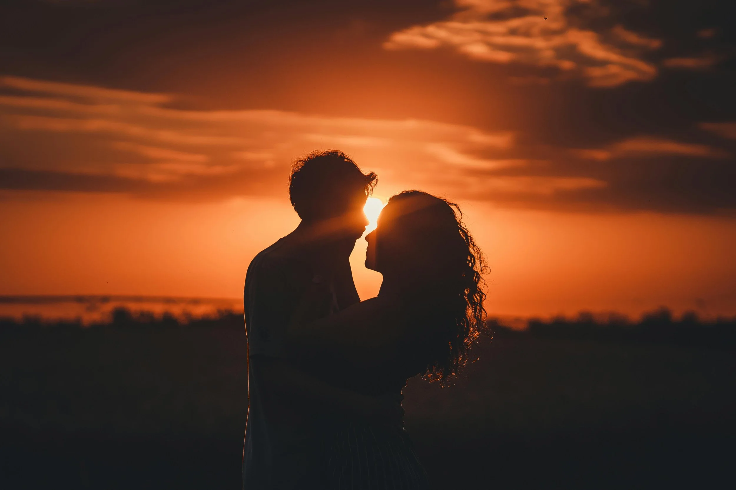 Una pareja en actitud romántica, abrazados y tocándose la frente, en un atardecer con cielo naranja y nubes oscuras.