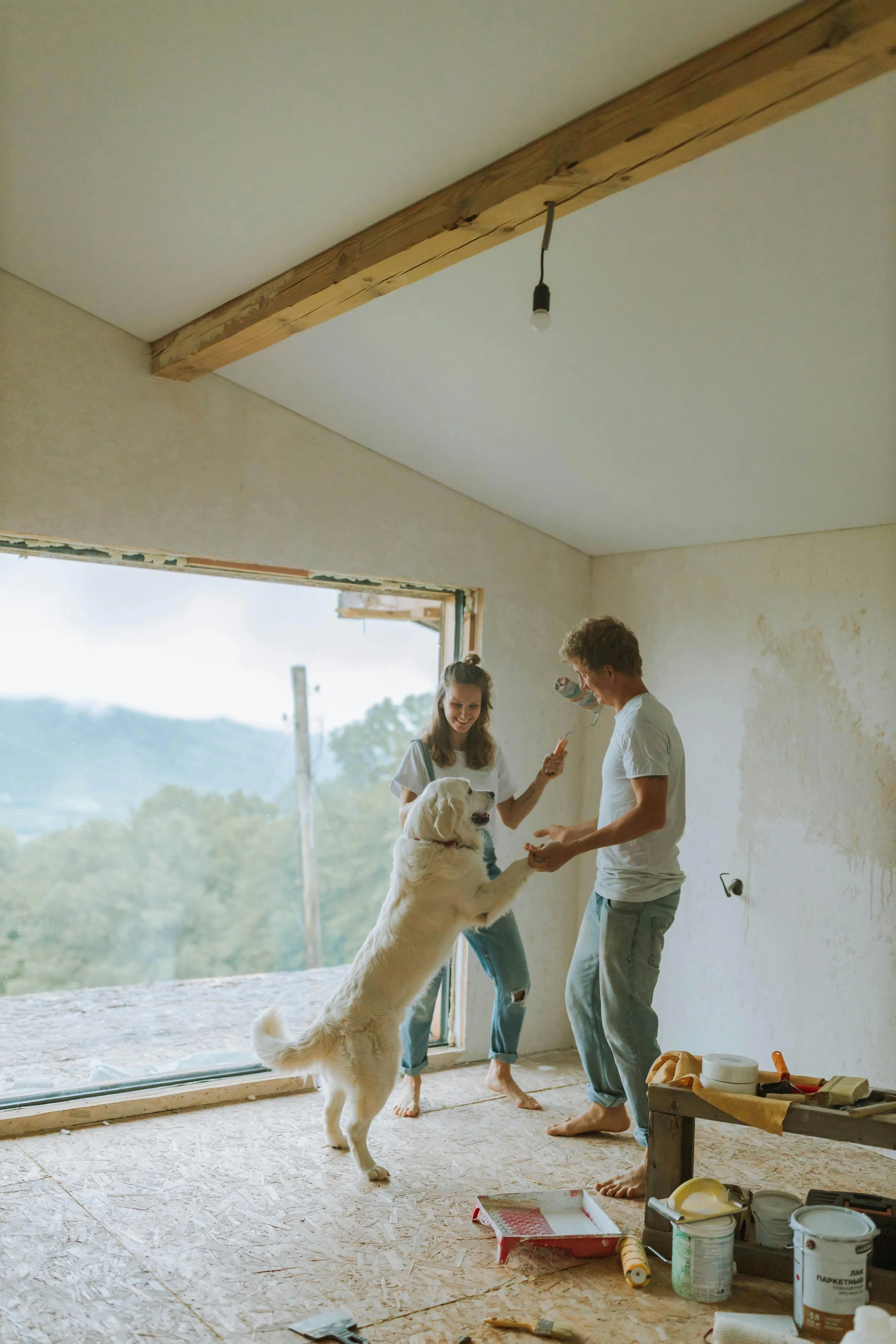 Una pareja y un perro en una habitación en construcción, con paredes sin terminar y herramientas de construcción en el suelo, disfrutando de un momento alegre.