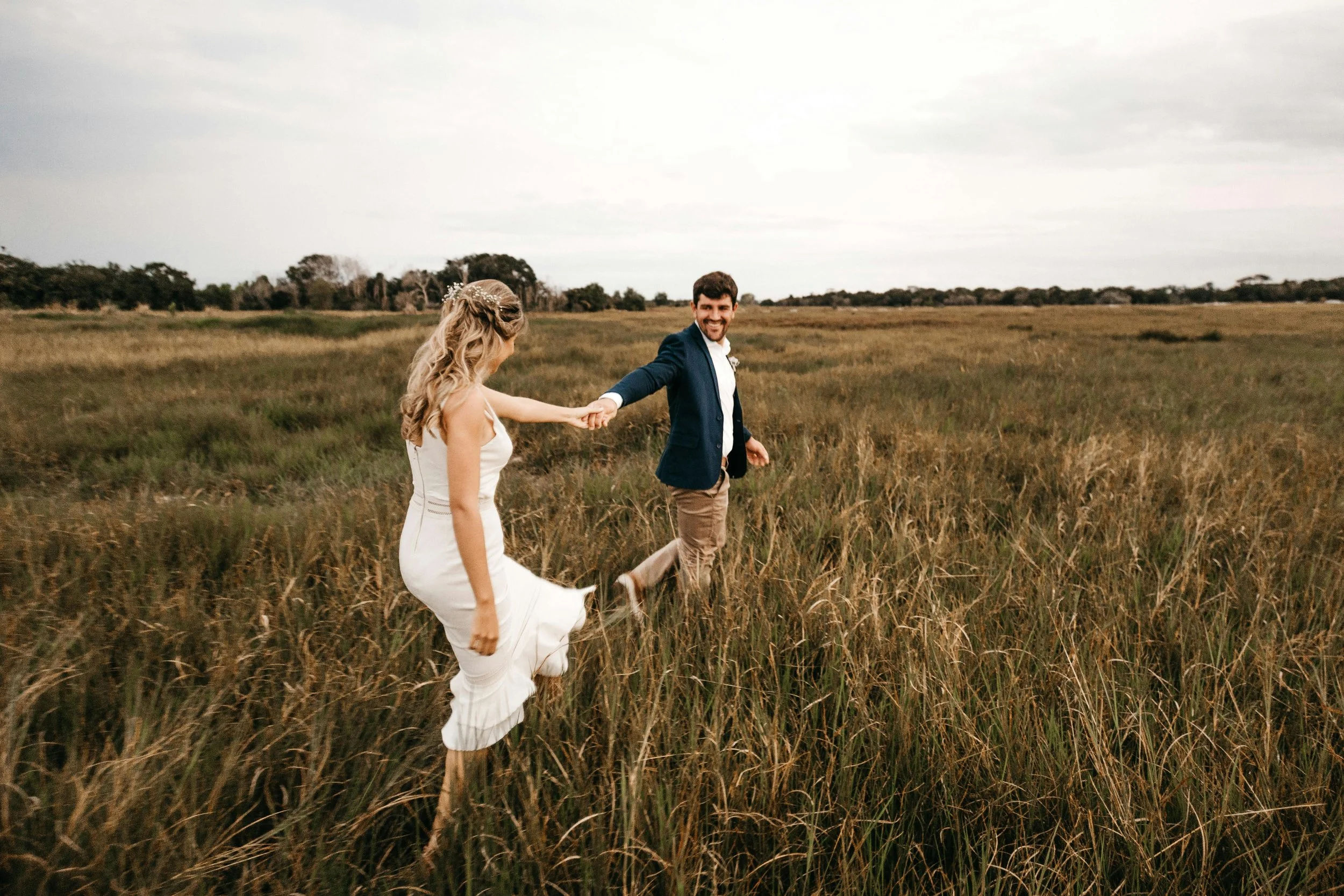 Pareja caminando de la mano en un campo abierto, ella lleva un vestido blanco y él lleva chaqueta azul y pantalones beige, parecen ser una pareja en una sesión de fotos al aire libre.