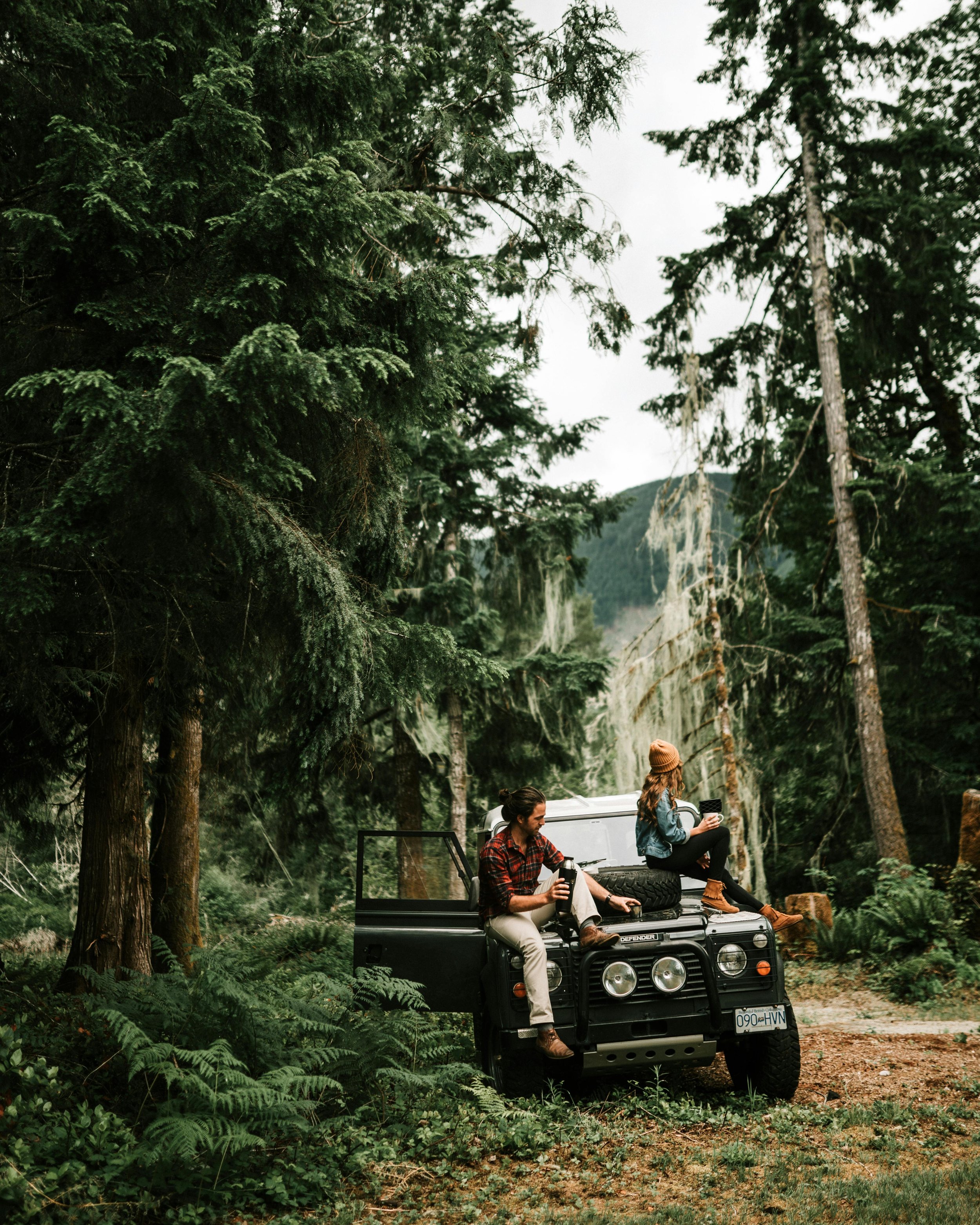 Un hombre y una mujer sentados en la cubierta de un vehículo todoterreno en un bosque, disfrutando de bebidas calientes, rodeados de árboles altos y vegetación densa.