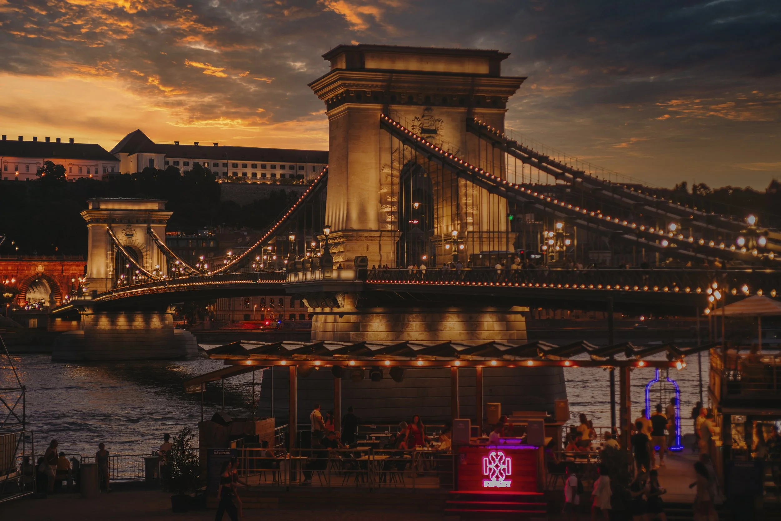 The Chain Bridge in Budapest illuminated at sunset with people gathered near the riverbank and a bar area in the foreground.