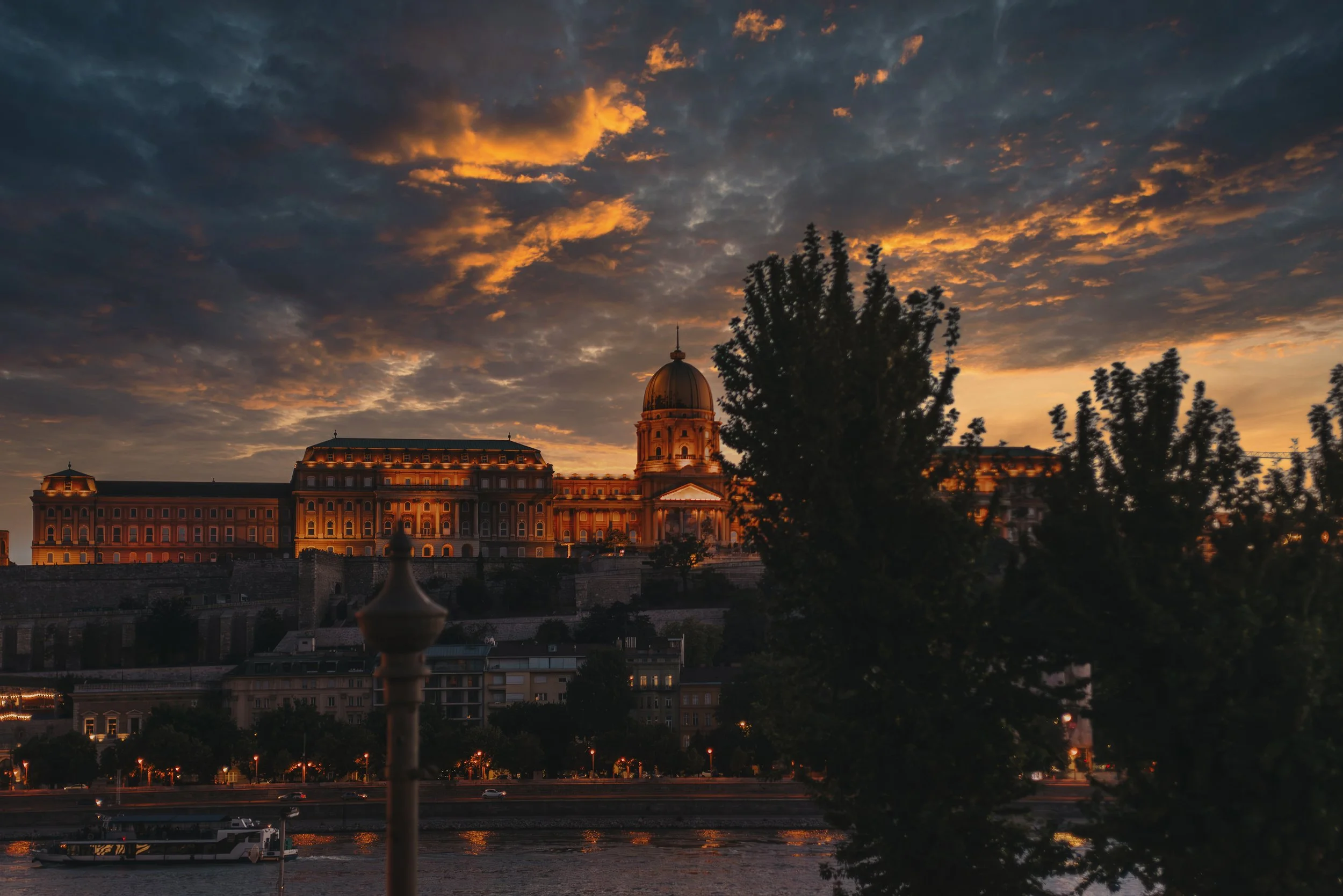 Sunset over a historic building with a dome, trees in the foreground, and a river below.