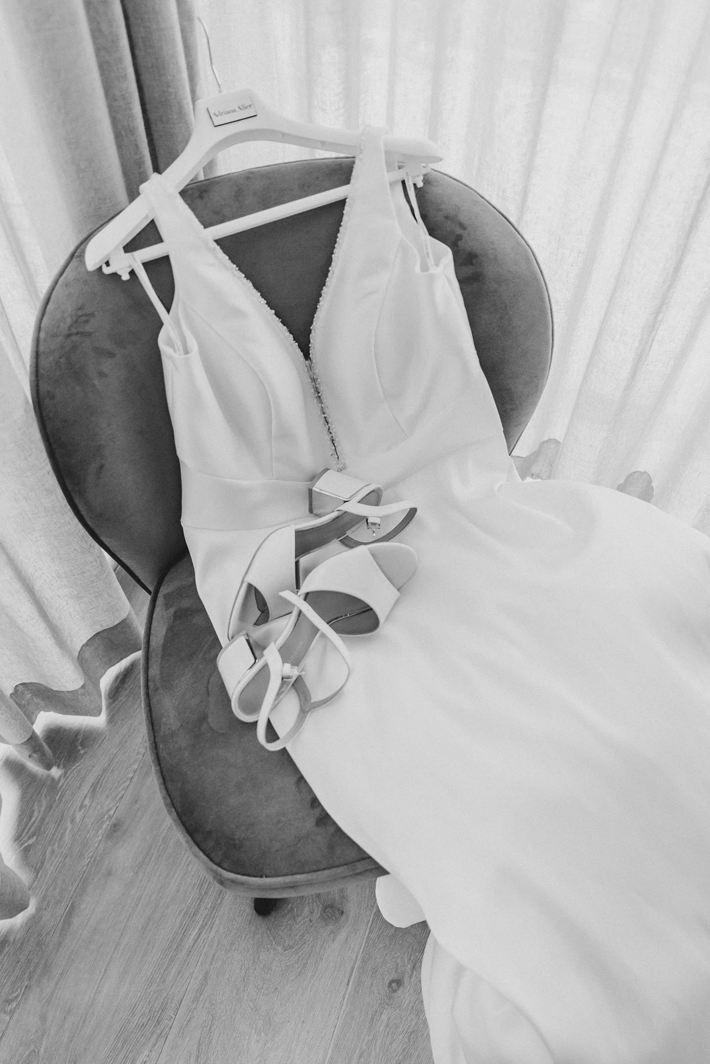 Vestido de novia blanco, con tacones y cinturón, colocados sobre una silla de terciopelo en una habitación con cortinas.