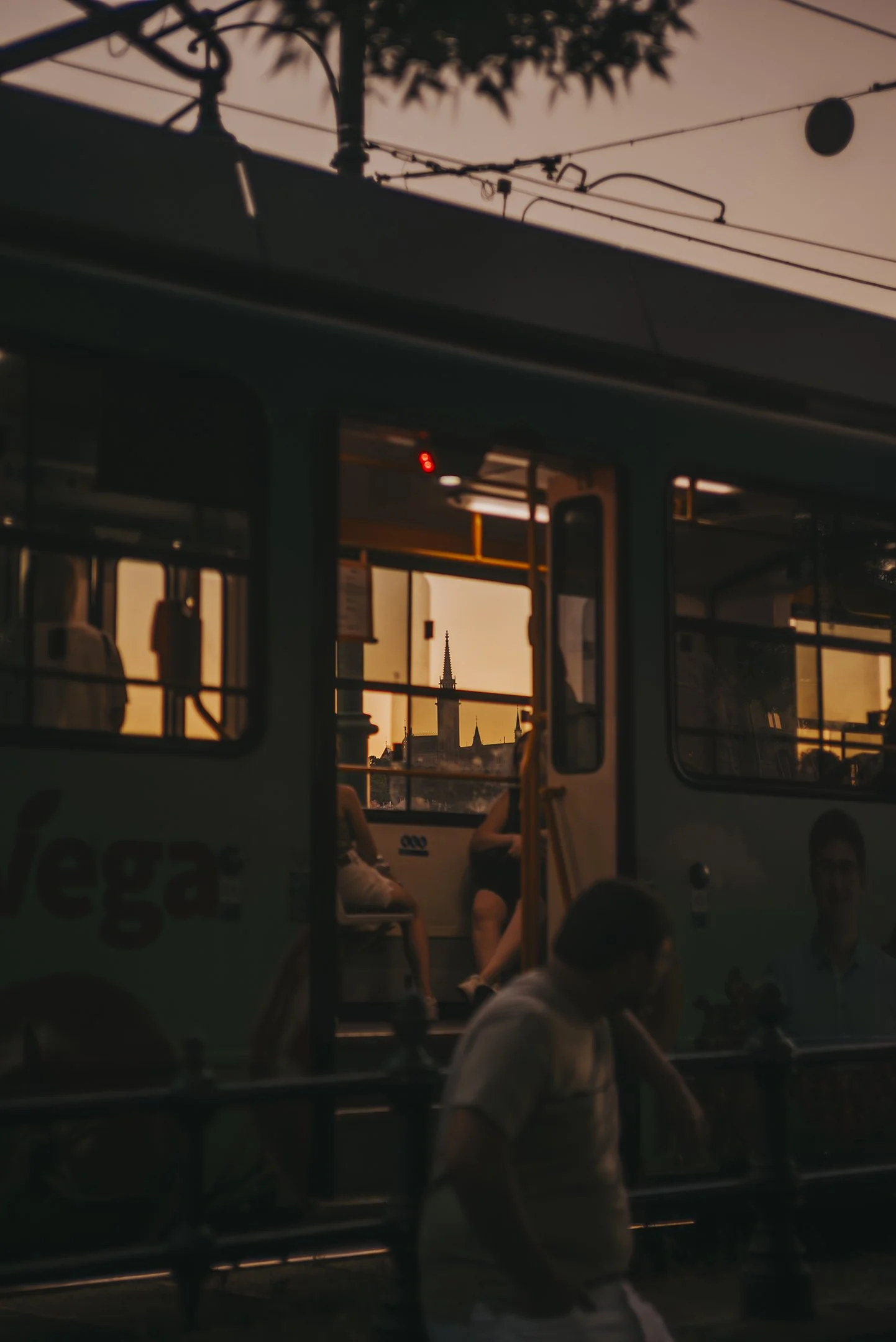 A city bus at sunset with silhouettes of passengers inside and a person outside in the foreground. A church steeple is visible through the bus window against a warm orange sky.