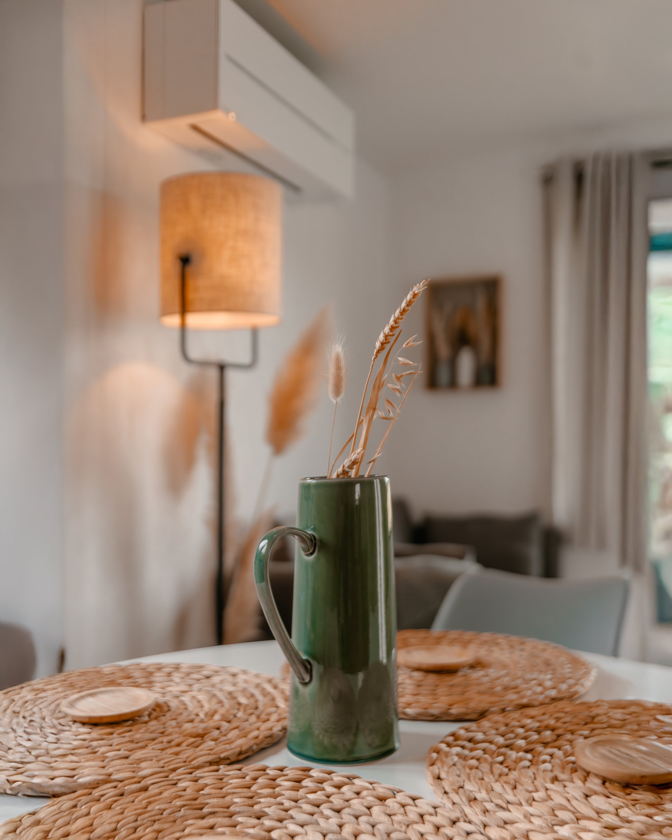 A dining table with a tall green vase holding dried grass on woven placemats, a wall-mounted lamp, and a blurry background of a living room with curtains and artwork.