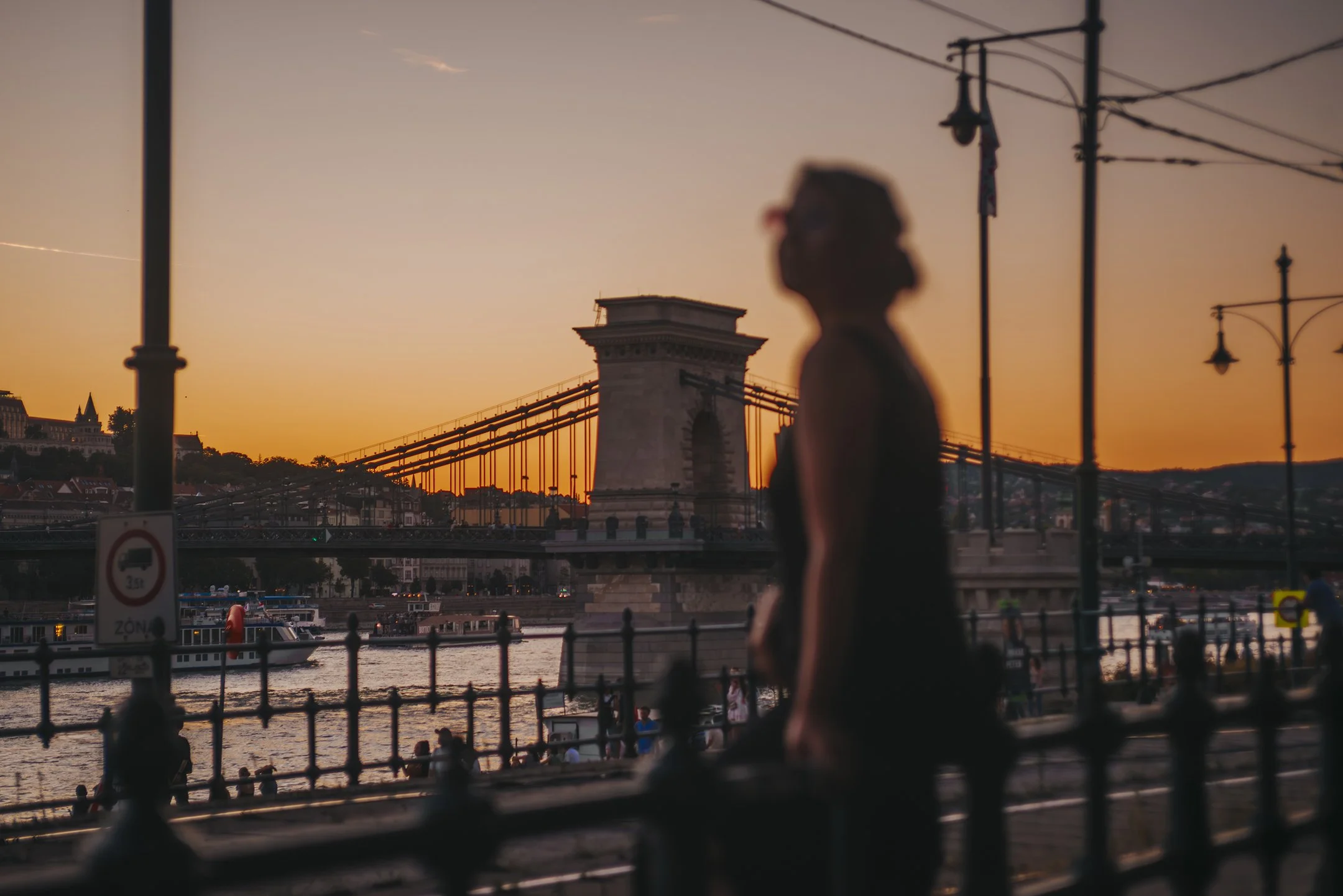 A woman stands near a river at sunset with the Chain Bridge and Buda Castle in the background, as boat cruise ships are on the water.