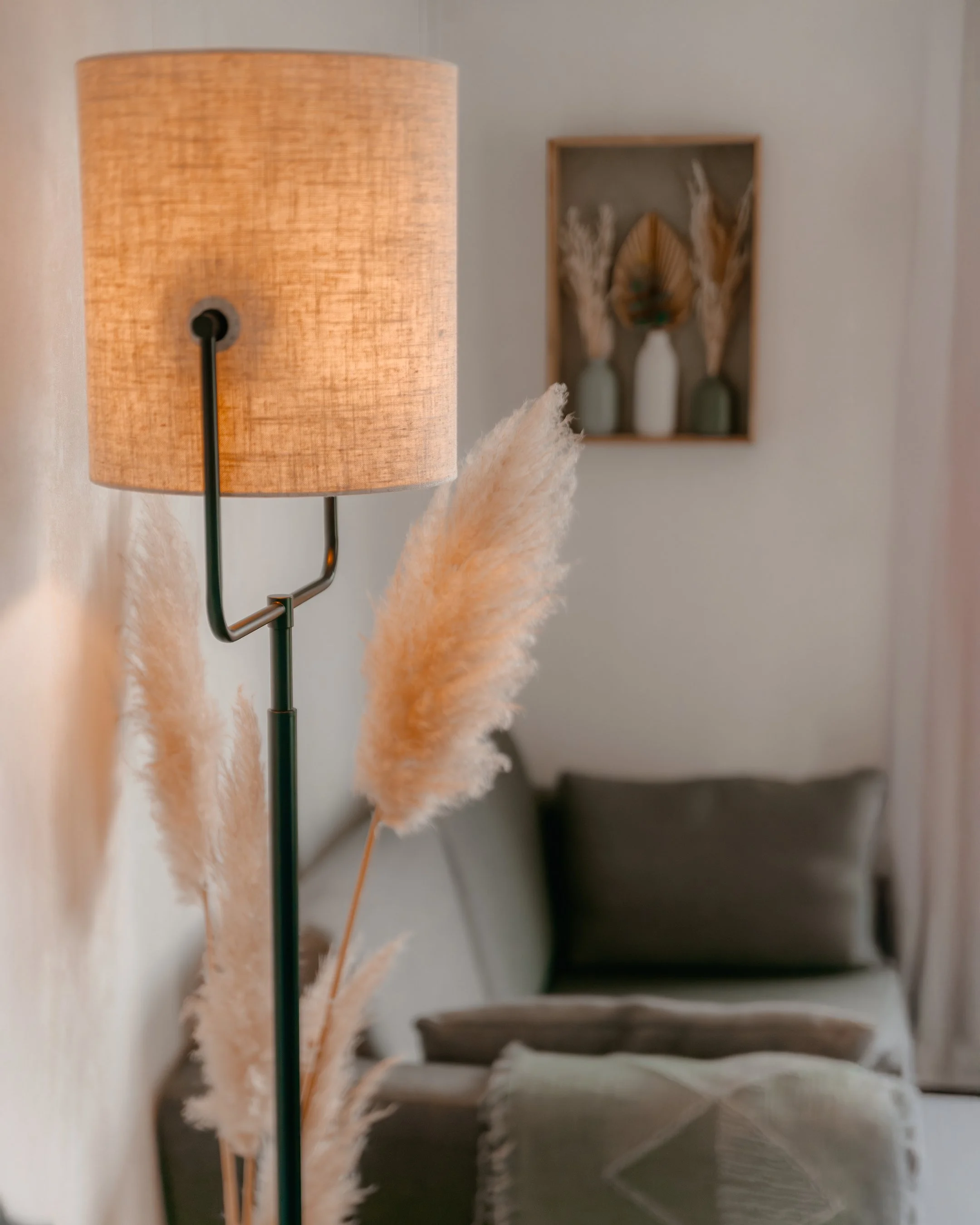 Close-up of a modern floor lamp with a beige lampshade, next to a bundle of dried pampas grass, in a living room with gray furniture and a wall decor featuring vases and dried plants.