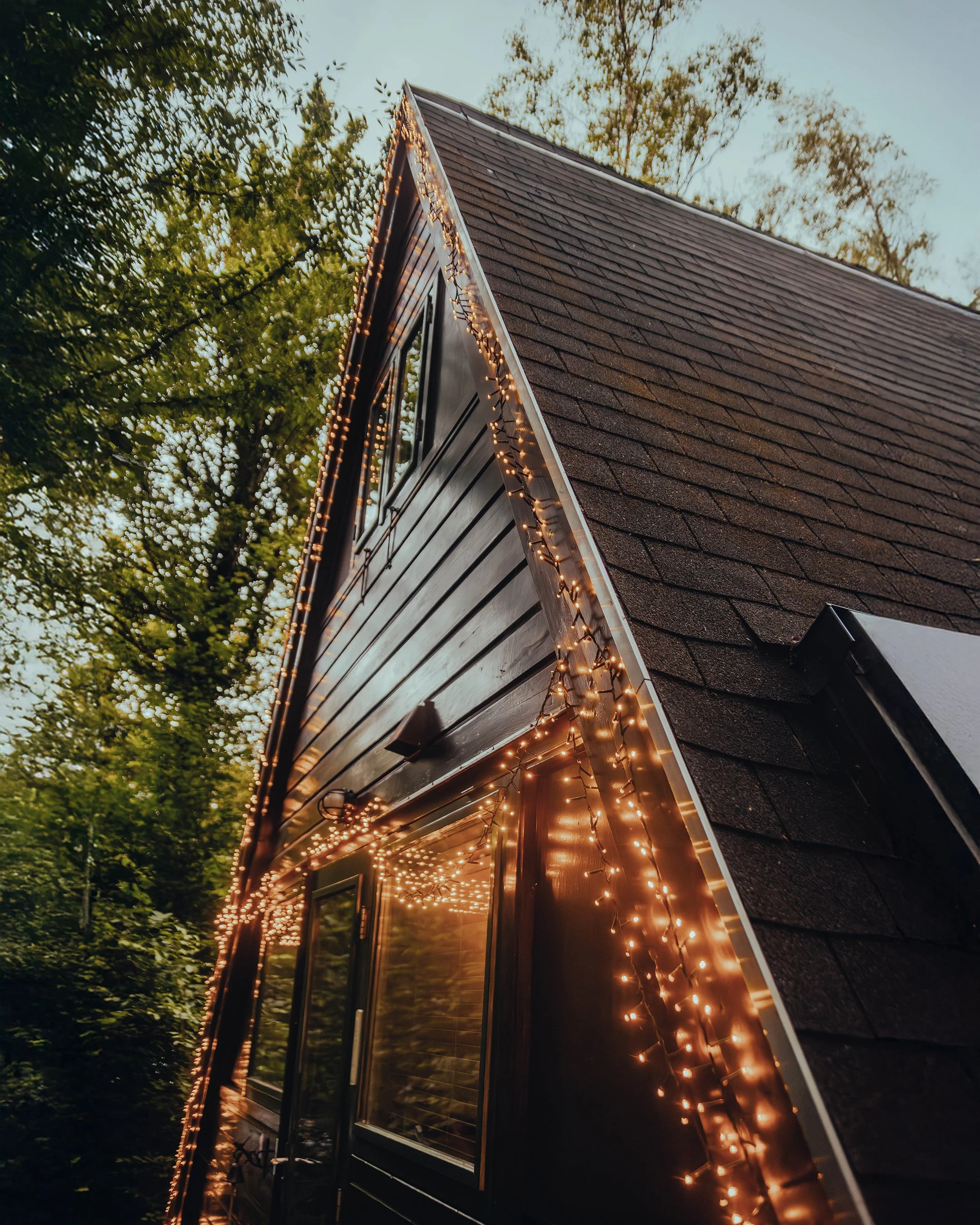 A house with fairy lights hanging along the roofline and around the window, with a dark wooden exterior and a steep shingle roof, surrounded by trees.