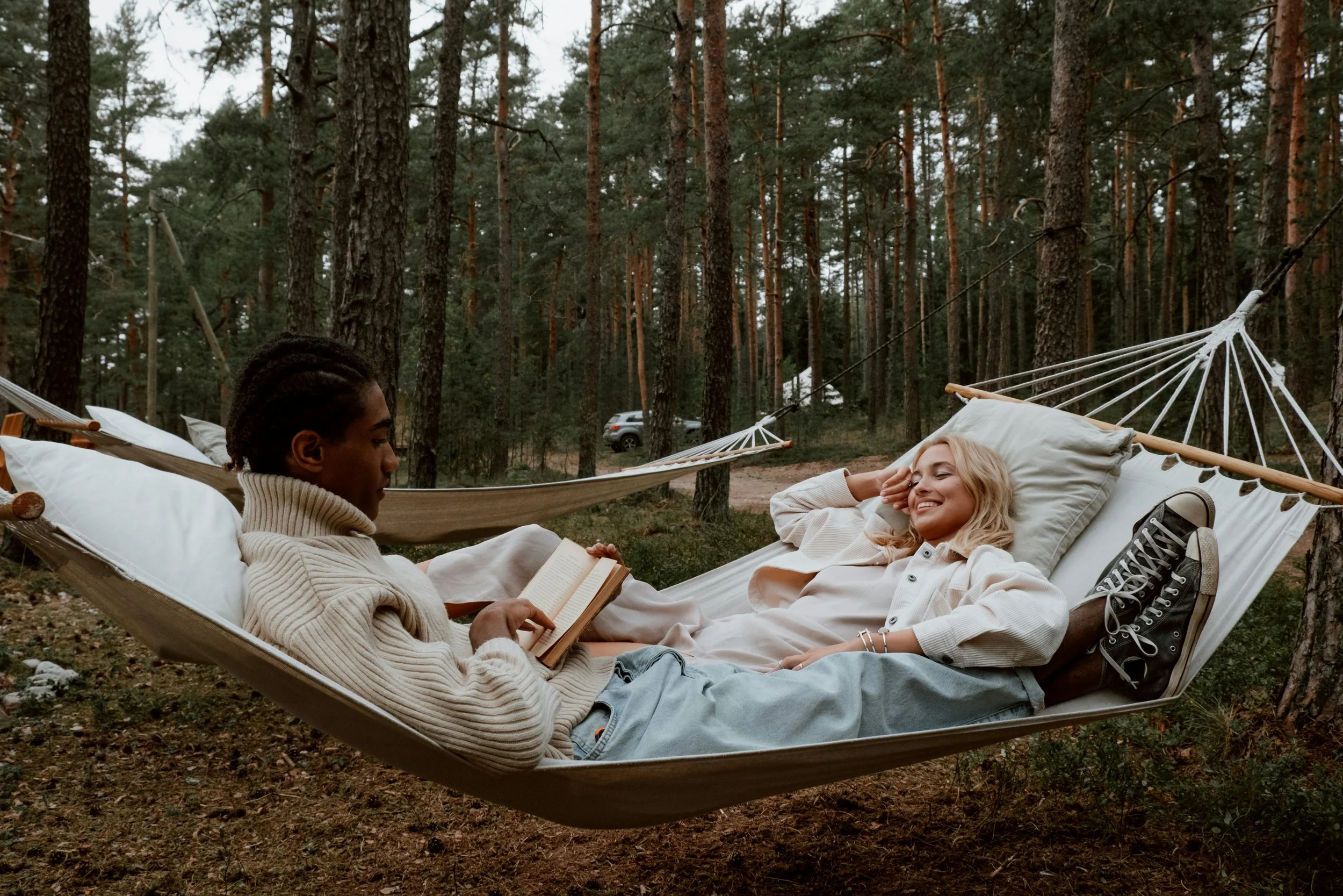 Dos personas descansando en hamacas en un bosque, una leyendo un libro y la otra sonriendo con los ojos cerrados.