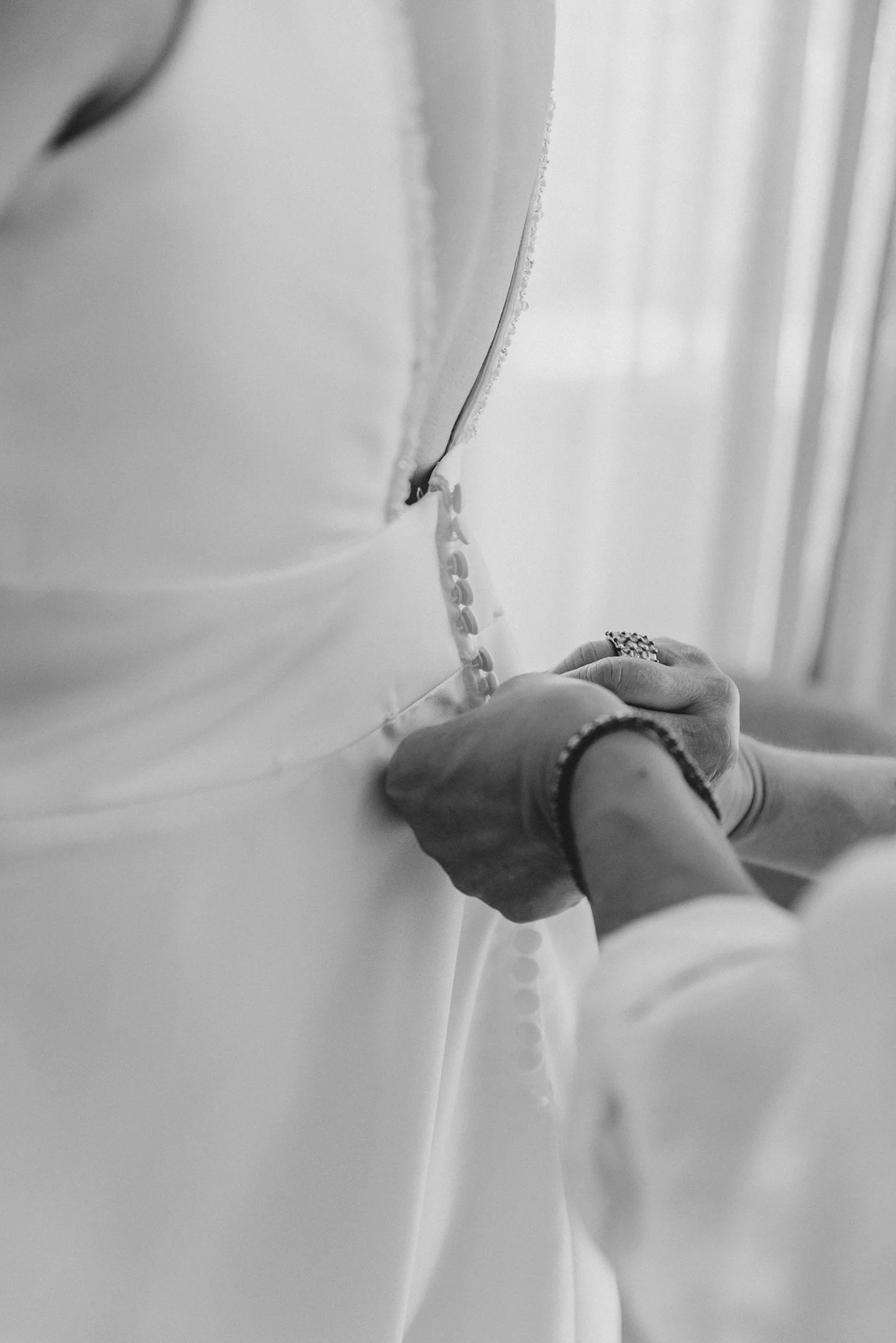 Mano cosiendo una prenda blanca con botones pequeños en un entorno interior, en blanco y negro.
