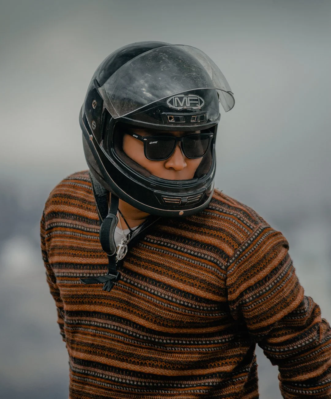 A person wearing a black motorcycle helmet, sunglasses, and a striped sweater.