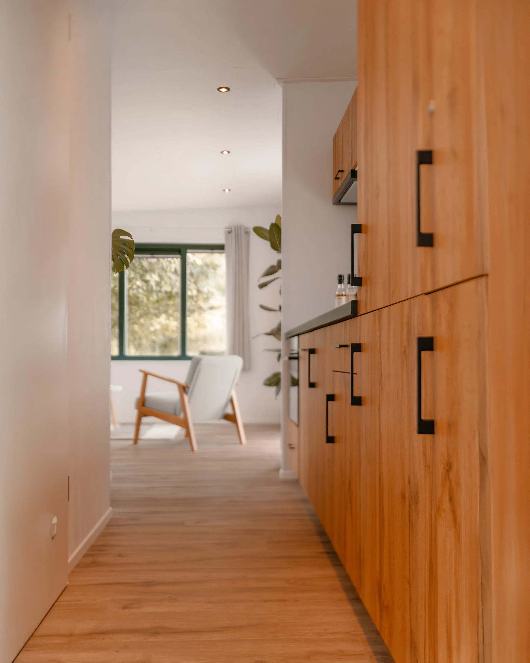 View through a doorway into a living room with wooden flooring, a large window with white curtains, a white armchair, and a potted plant.
