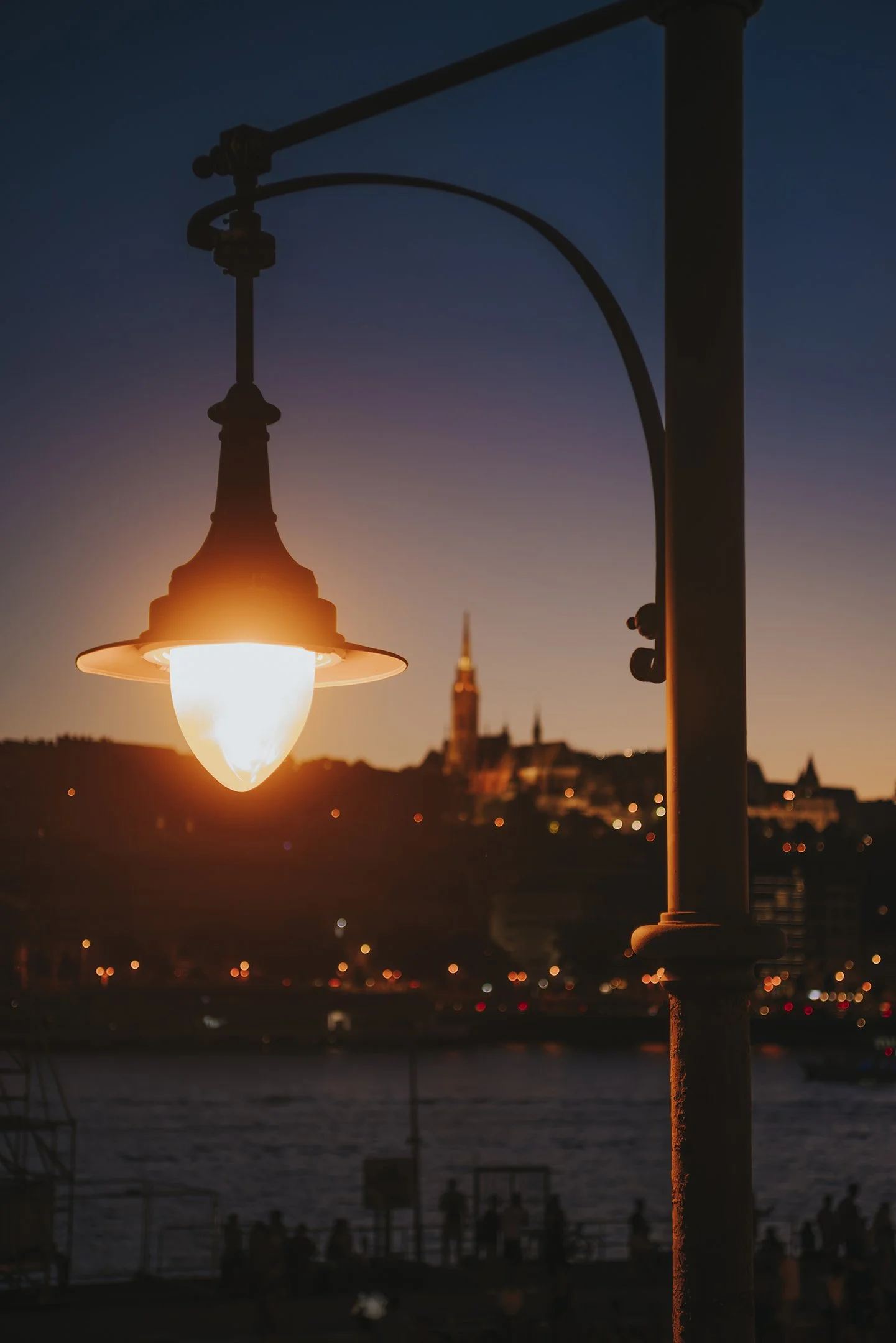 Close-up of a street lamp at sunset with a city skyline and a body of water in the background.