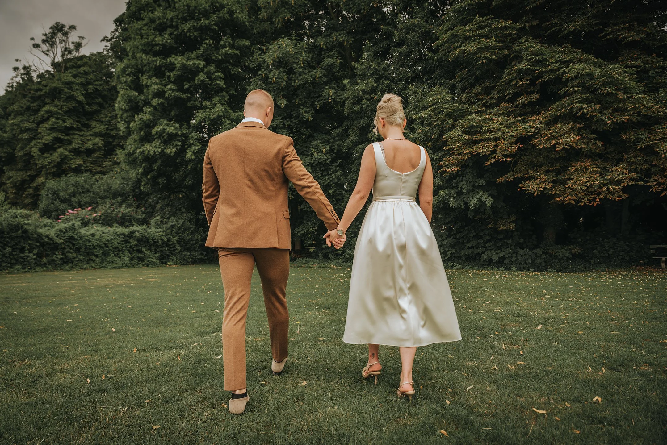 Pareja de novios caminando por el césped, vista desde atrás, con árboles en el fondo, ella lleva vestido blanco y él lleva traje marrón.