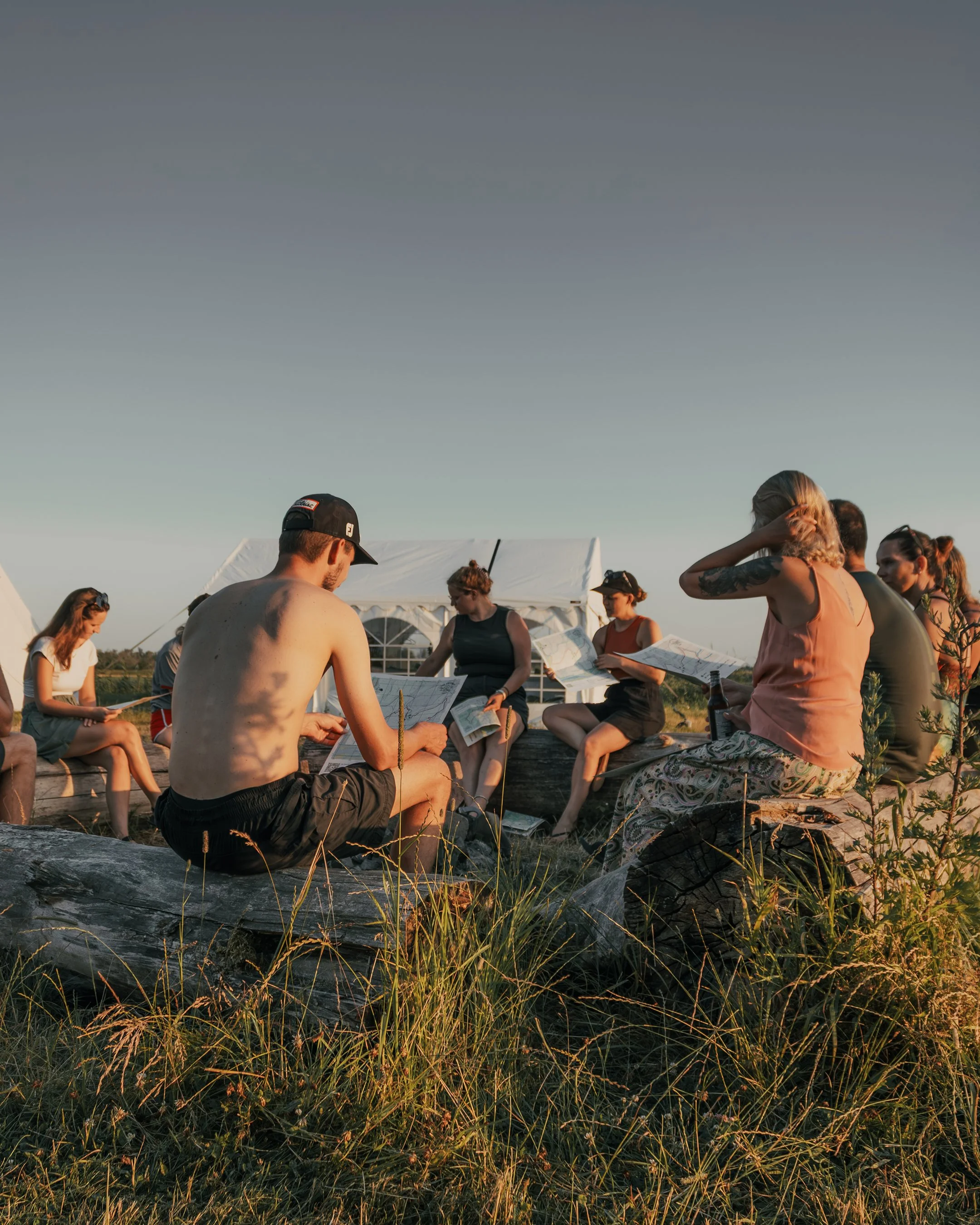 Group of people sitting outdoors near tents, reading papers, during sunset.