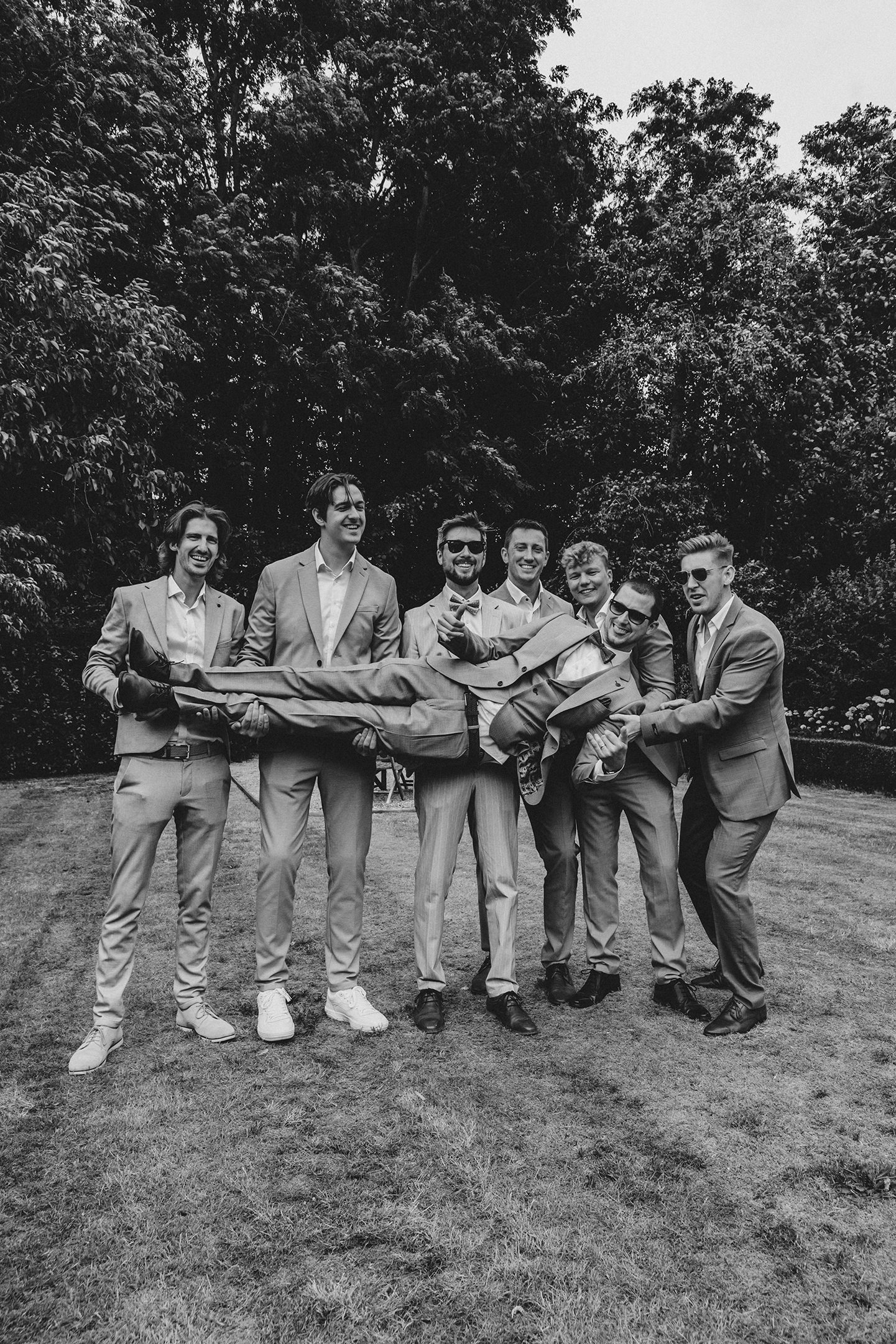 Grupo de siete hombres vestidos con trajes sosteniendo a un hombre en posición horizontal en un espacio al aire libre con árboles en el fondo, todos sonrientes.