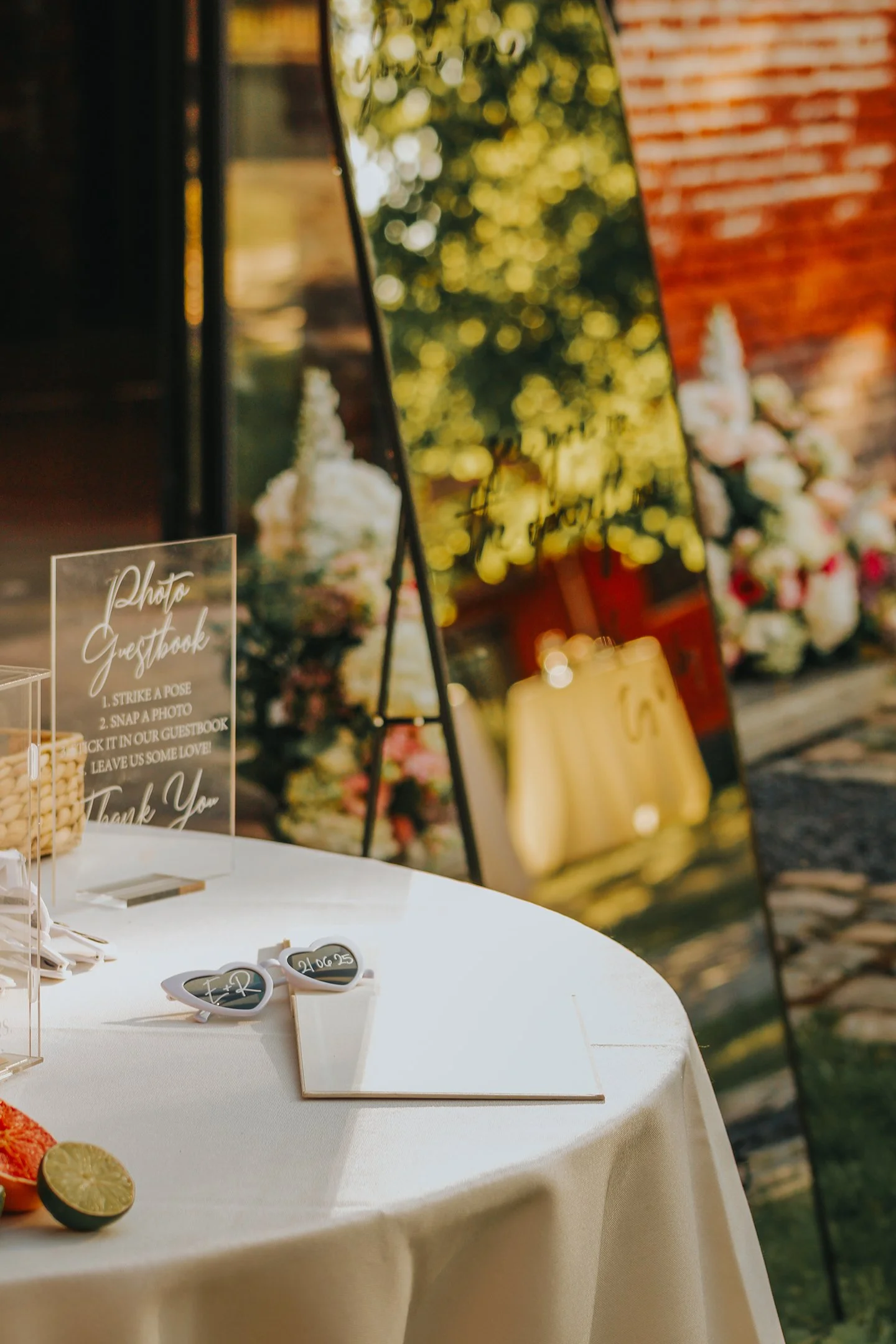 Mesa decorada para evento con gafas de sol y cartel que dice 'Photo Guestbook' con instrucciones para tomar fotos y dejar recuerdos en un libro de invitado, en un ambiente al aire libre con flores y espejos decorativos.