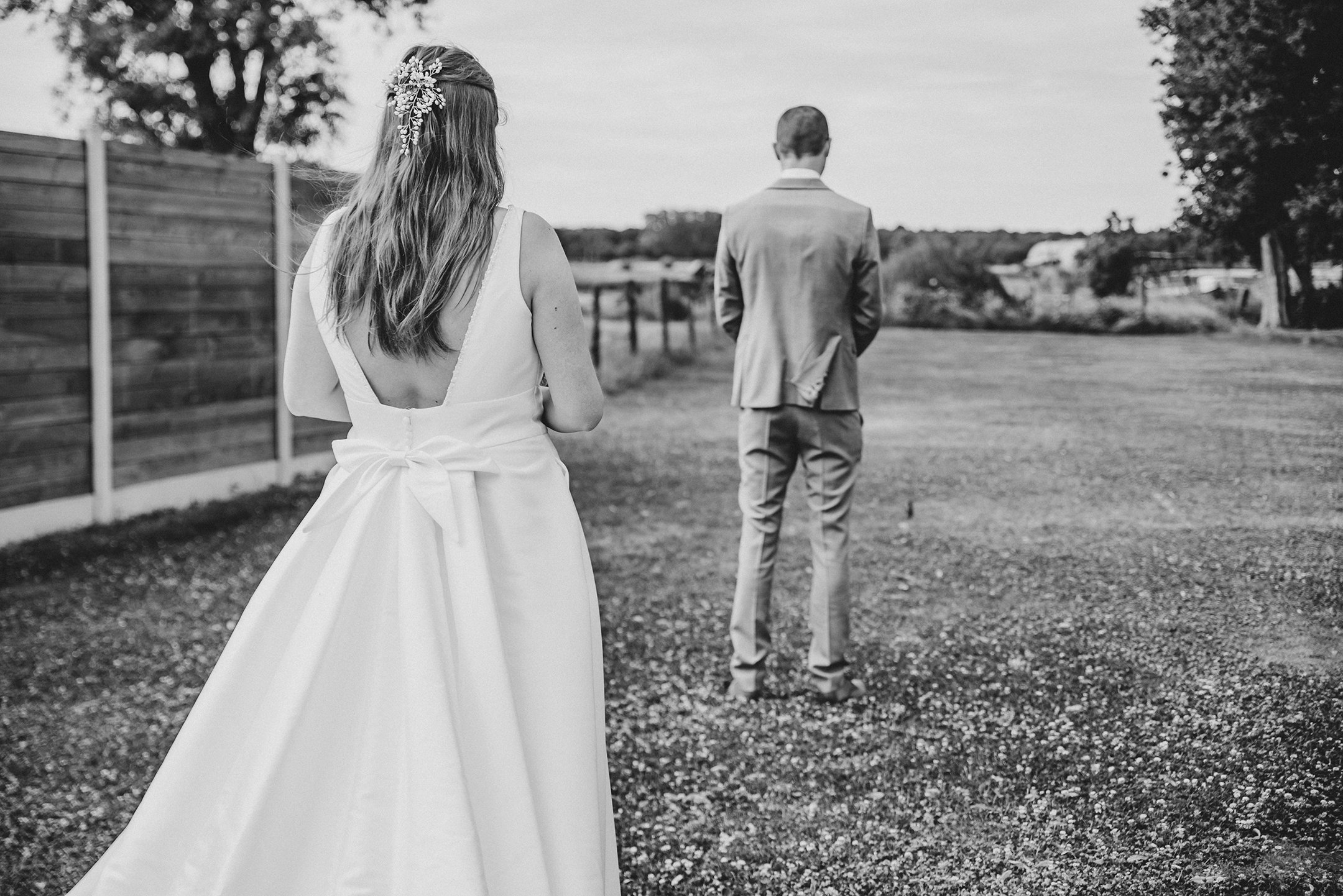 Una pareja vestida con ropa formal de boda caminando por un camino de tierra, con la mujer usando un vestido largo y un adorno en el cabello, la imagen en blanco y negro.